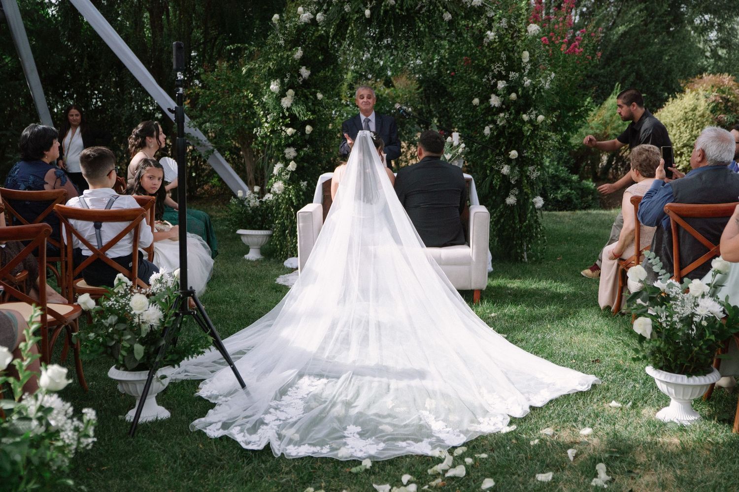 Bride with long cathedral veil standing at outdoor garden wedding altar with officiant.