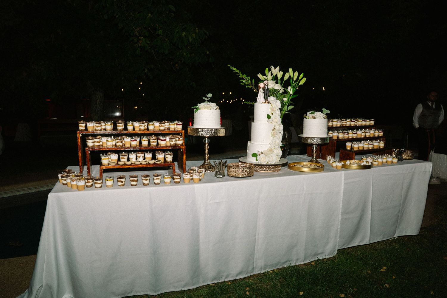 Elegant wedding dessert table with white cake and golden mini treats displayed outdoors at night.