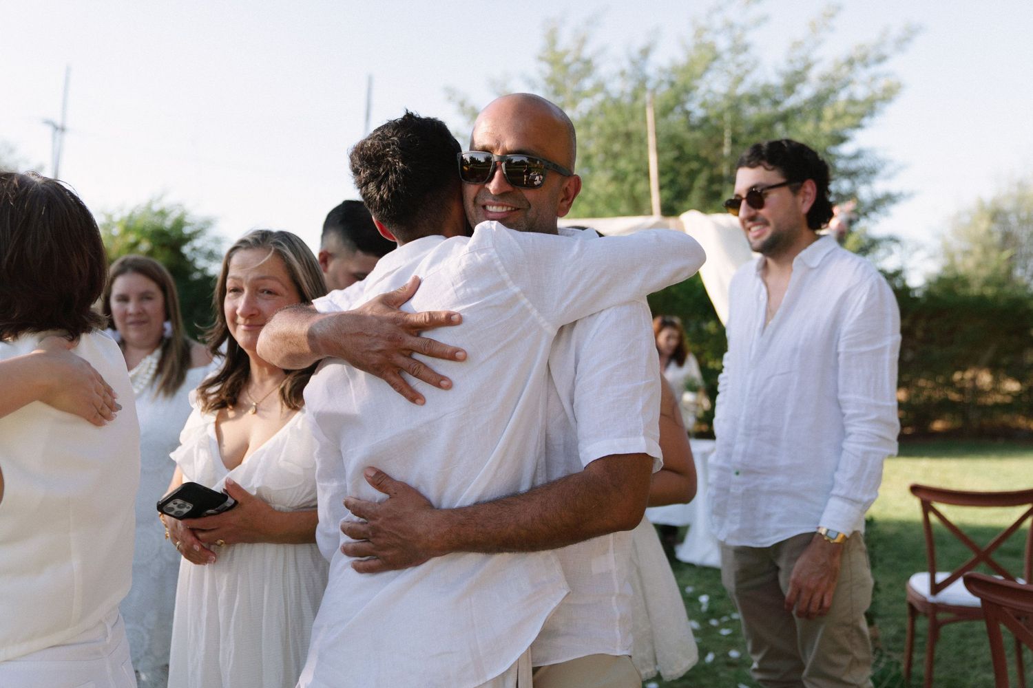 Wedding guests share warm embraces and congratulations outdoors following a garden wedding ceremony.