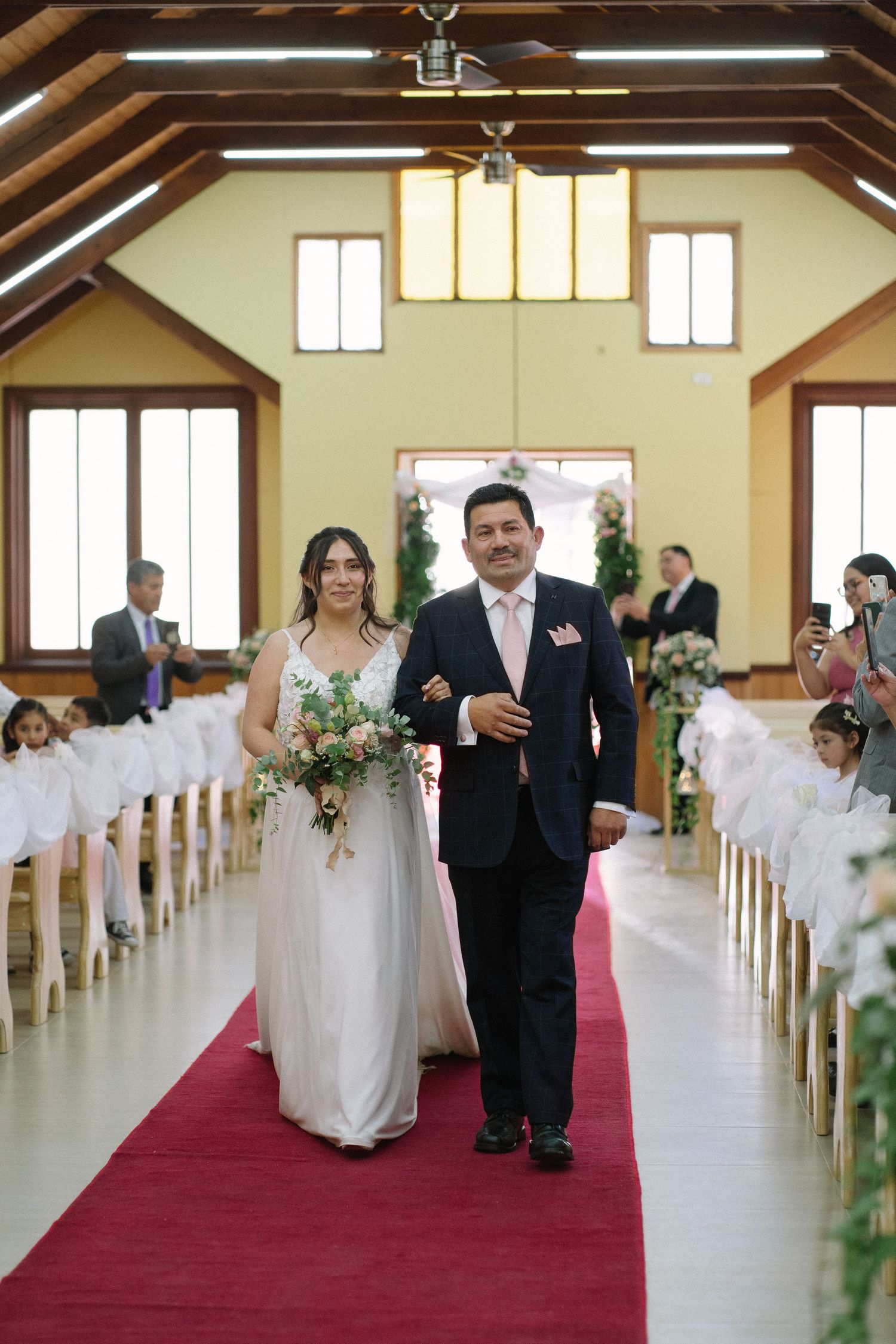A wedding processional walks down a red-carpeted aisle in a church with wooden beam architecture.