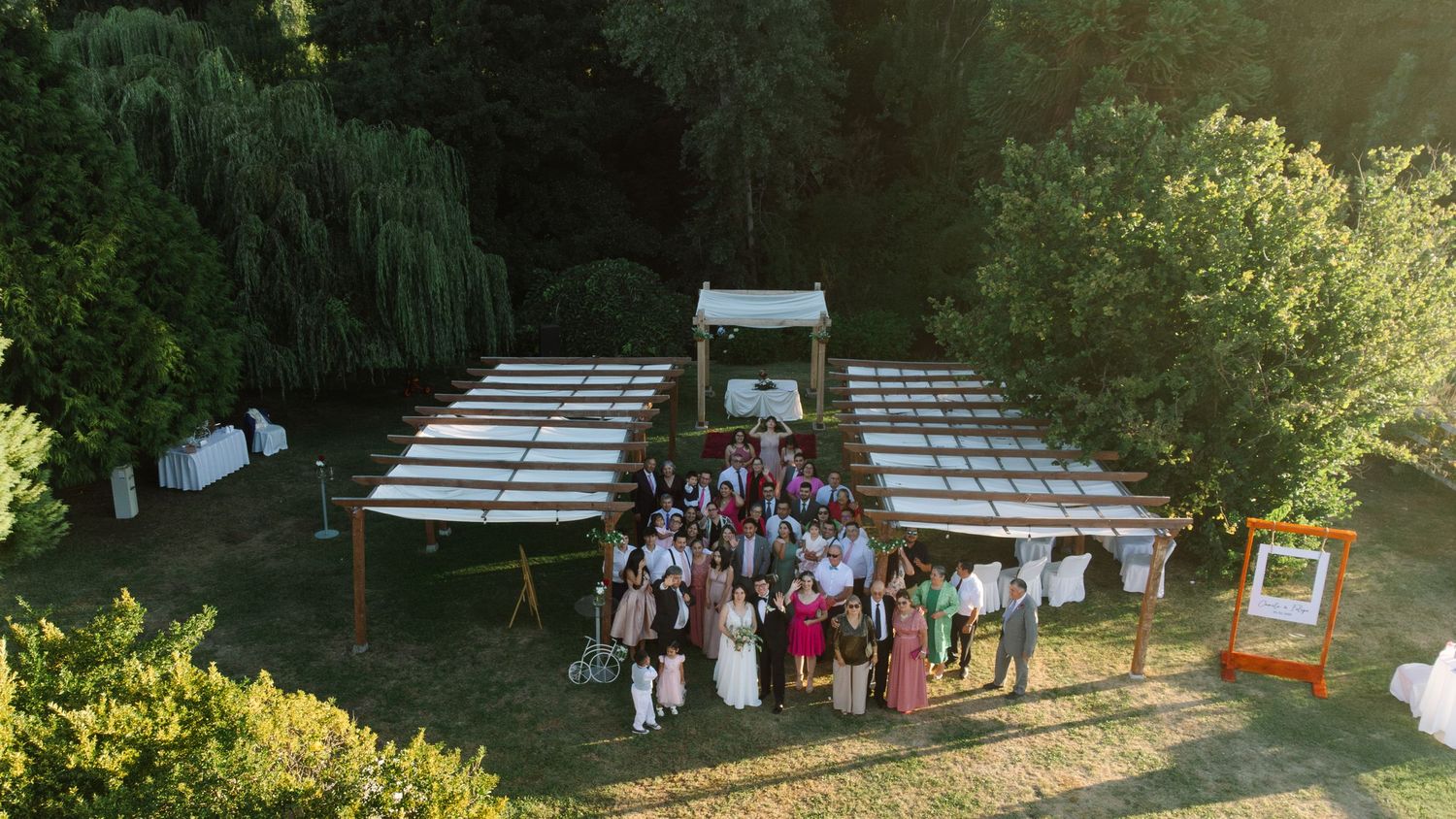 Aerial view of an outdoor wedding ceremony setup with white chairs and guests gathered under trees.