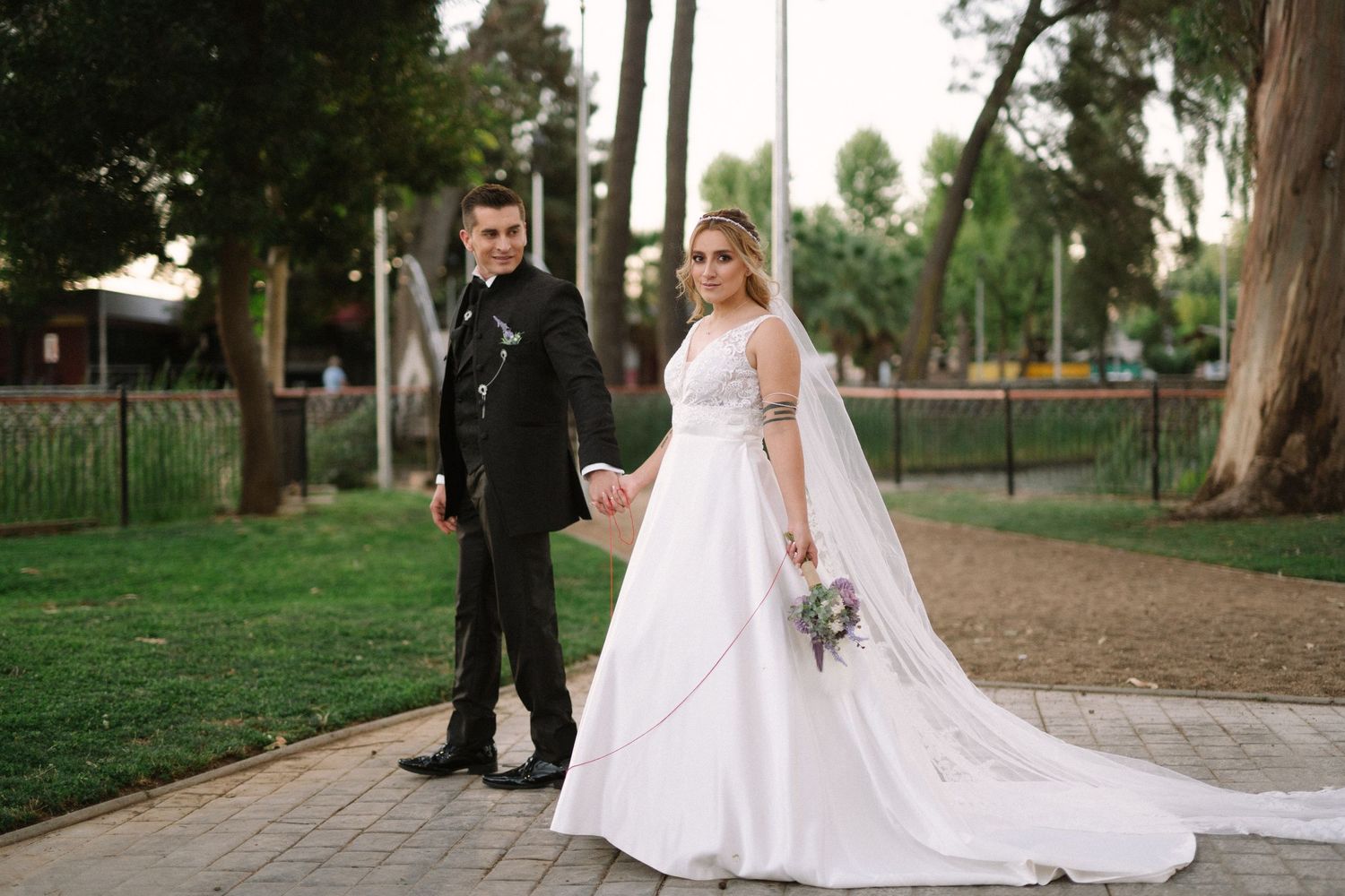 A wedding couple poses on a pathway with trees and iron fencing in the background.