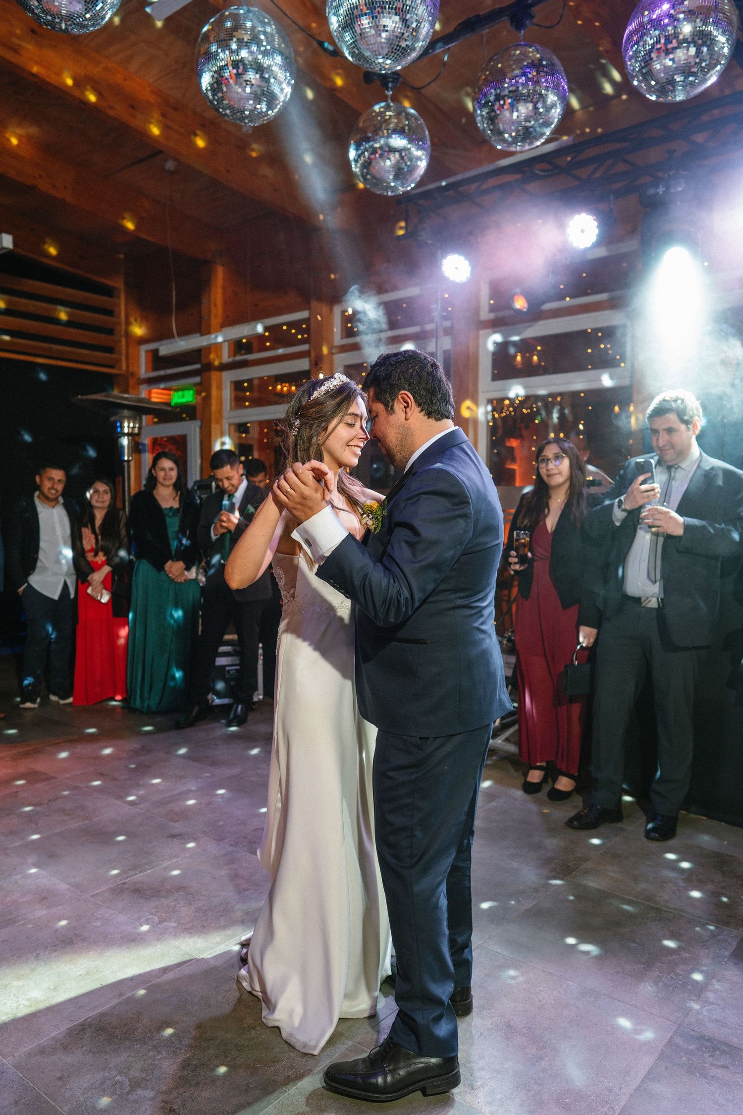 A couple shares their first dance under disco balls at a wedding reception surrounded by guests.
