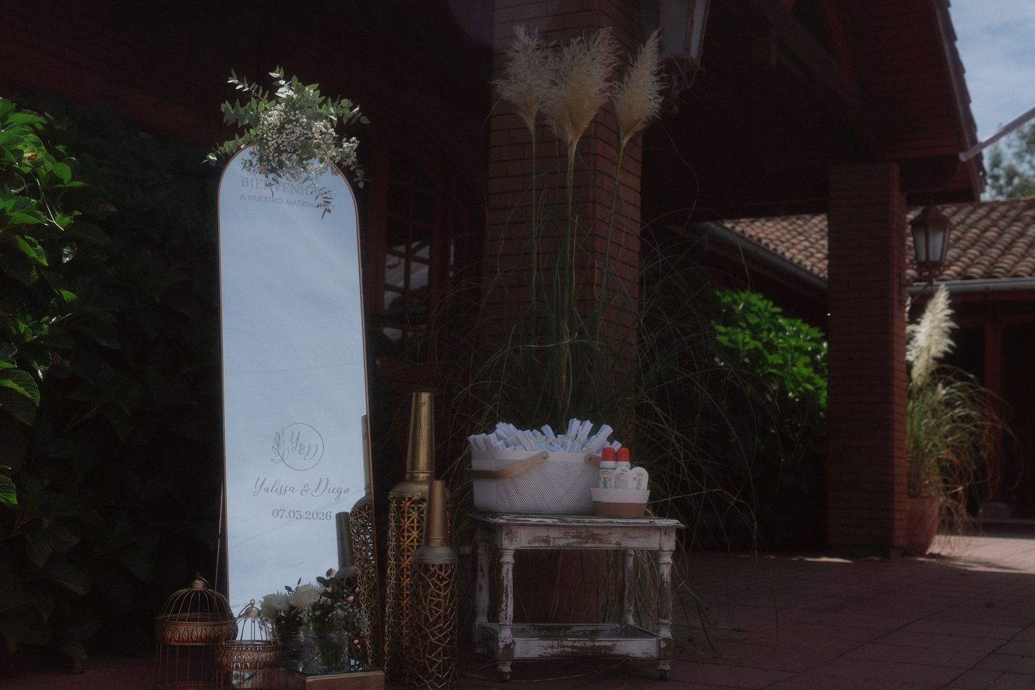 Elegant outdoor wedding display with white mirror, pampas grass, and rustic white table under a wooden pergola.
