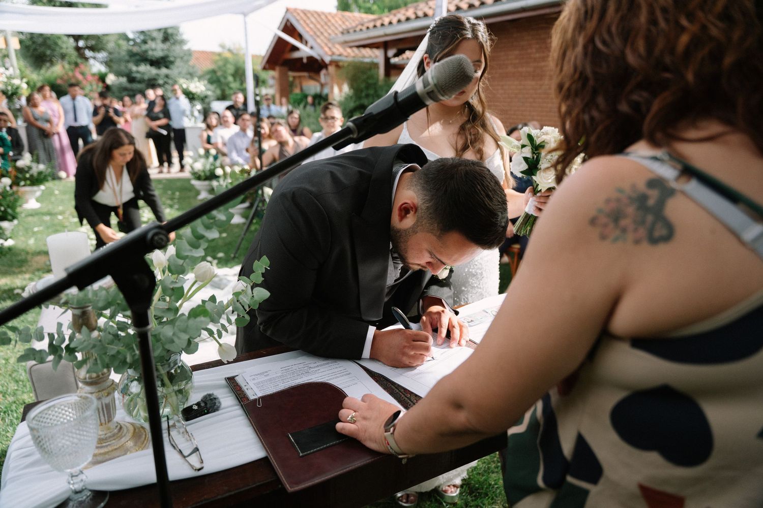 Groom signing wedding registry documents at outdoor ceremony table with guests in background.