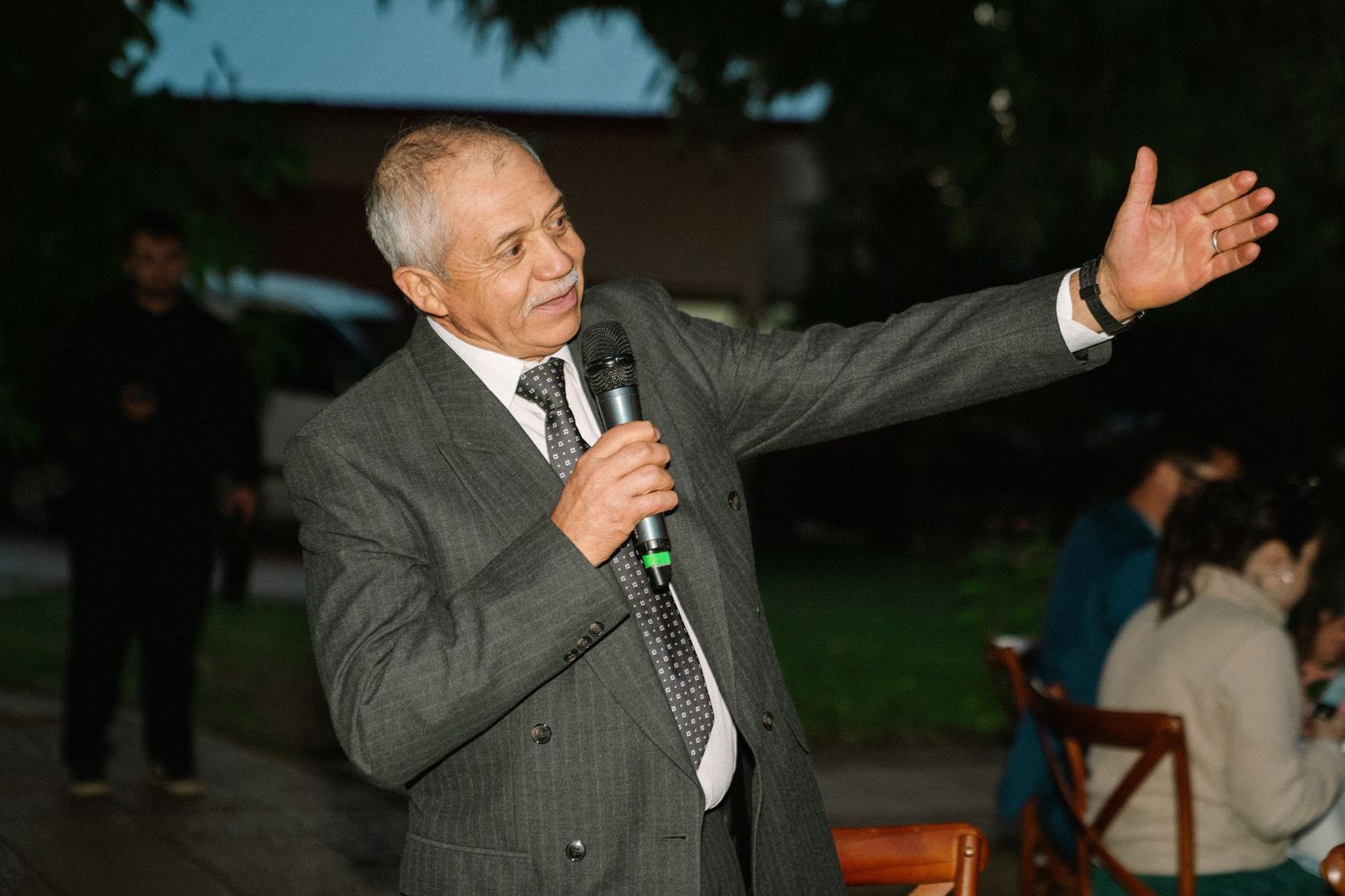 Elderly man in gray suit speaking into microphone with hand raised at outdoor wedding.