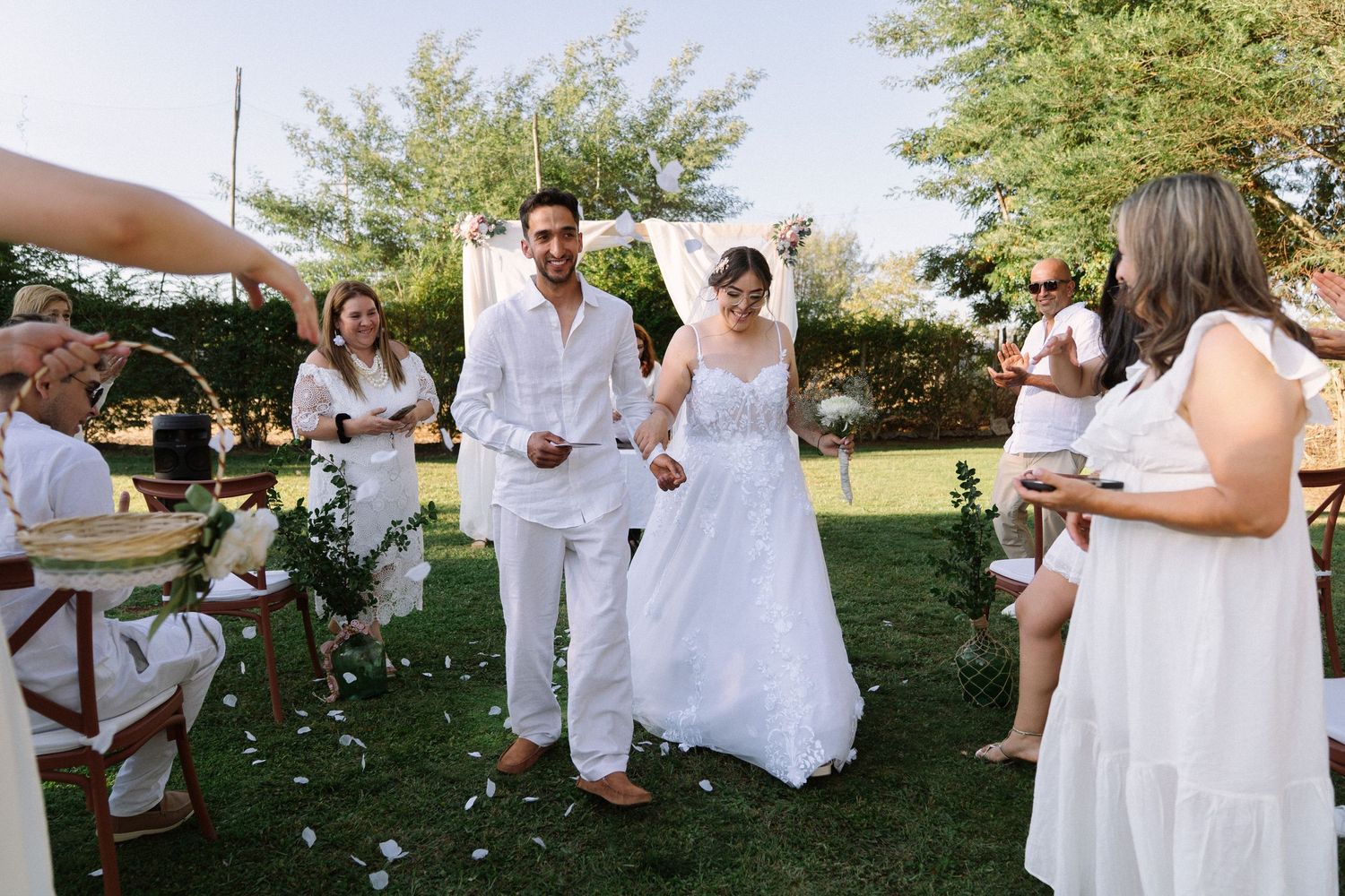 Bride and groom exit outdoor wedding ceremony as guests toss rose petals under sunny sky.