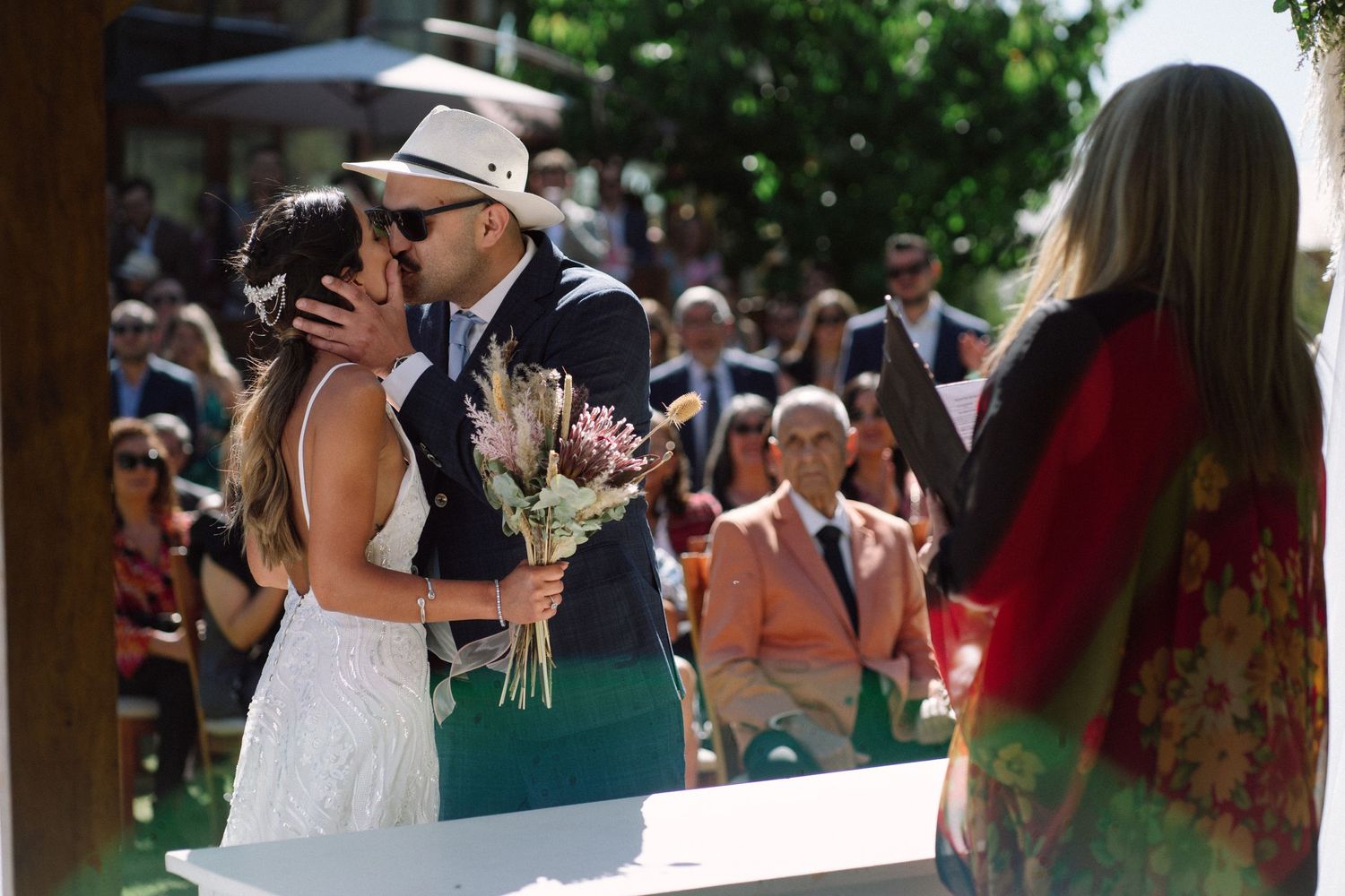 Outdoor wedding ceremony under natural light with gathered guests in background.