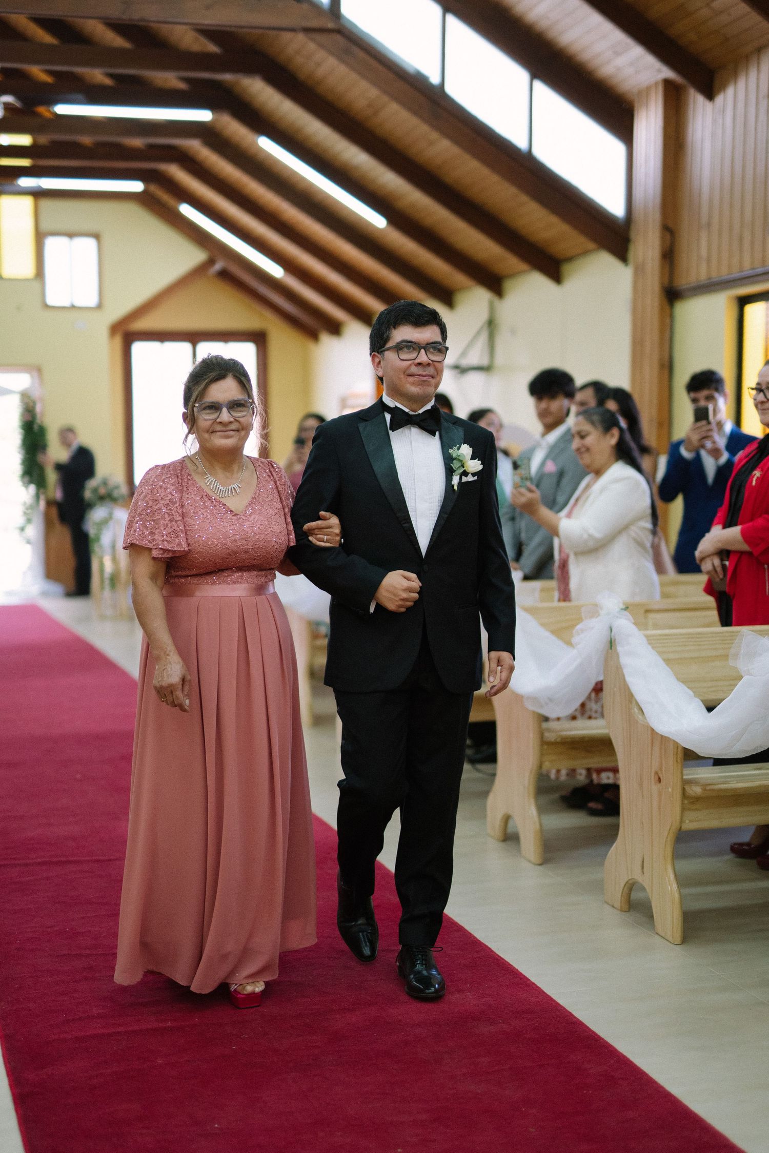 A procession walks down a red-carpeted church aisle during a formal ceremony with wooden beam ceilings.
