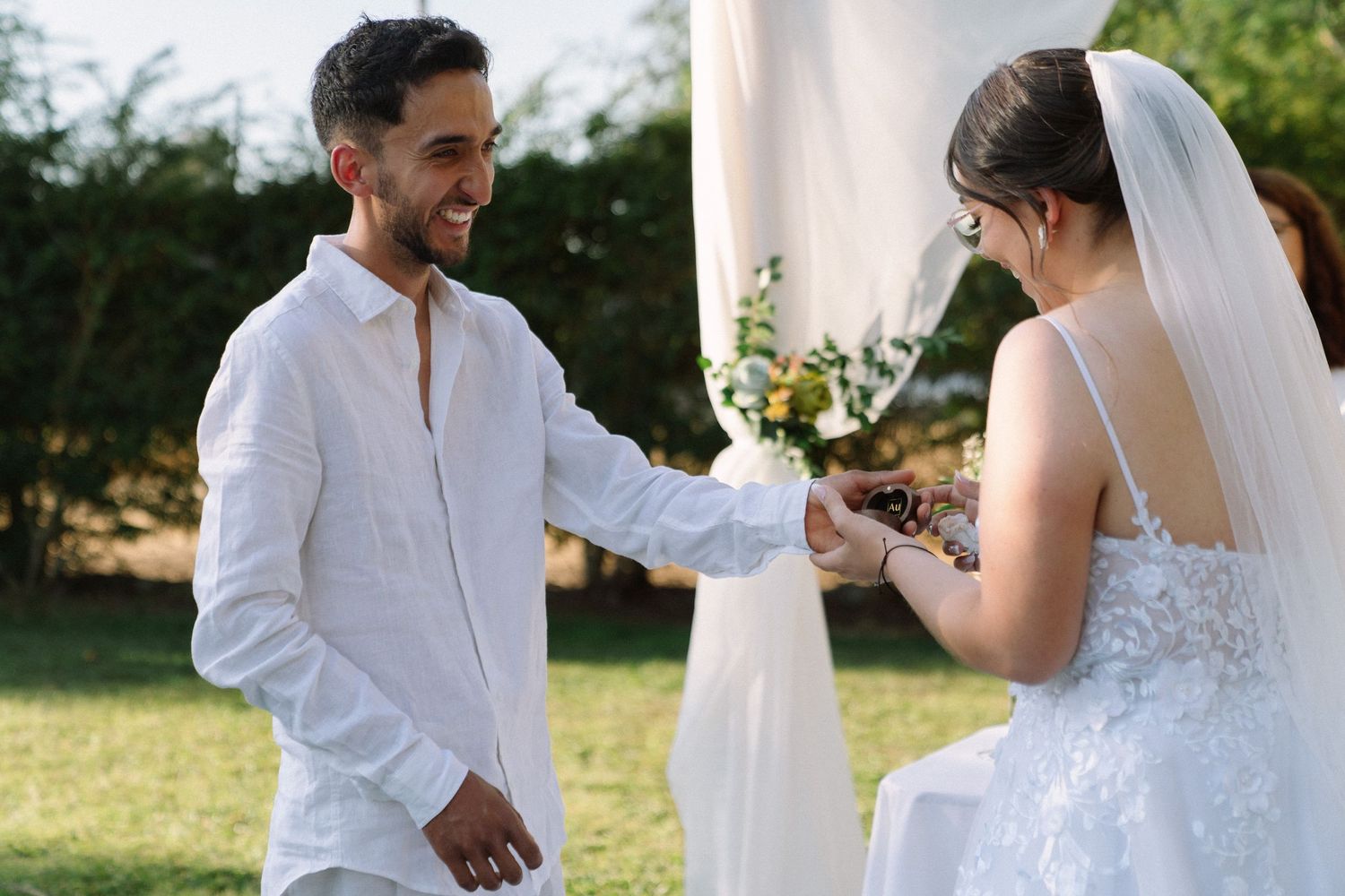 Smiling groom holds bride's hands during outdoor wedding ring exchange ceremony.