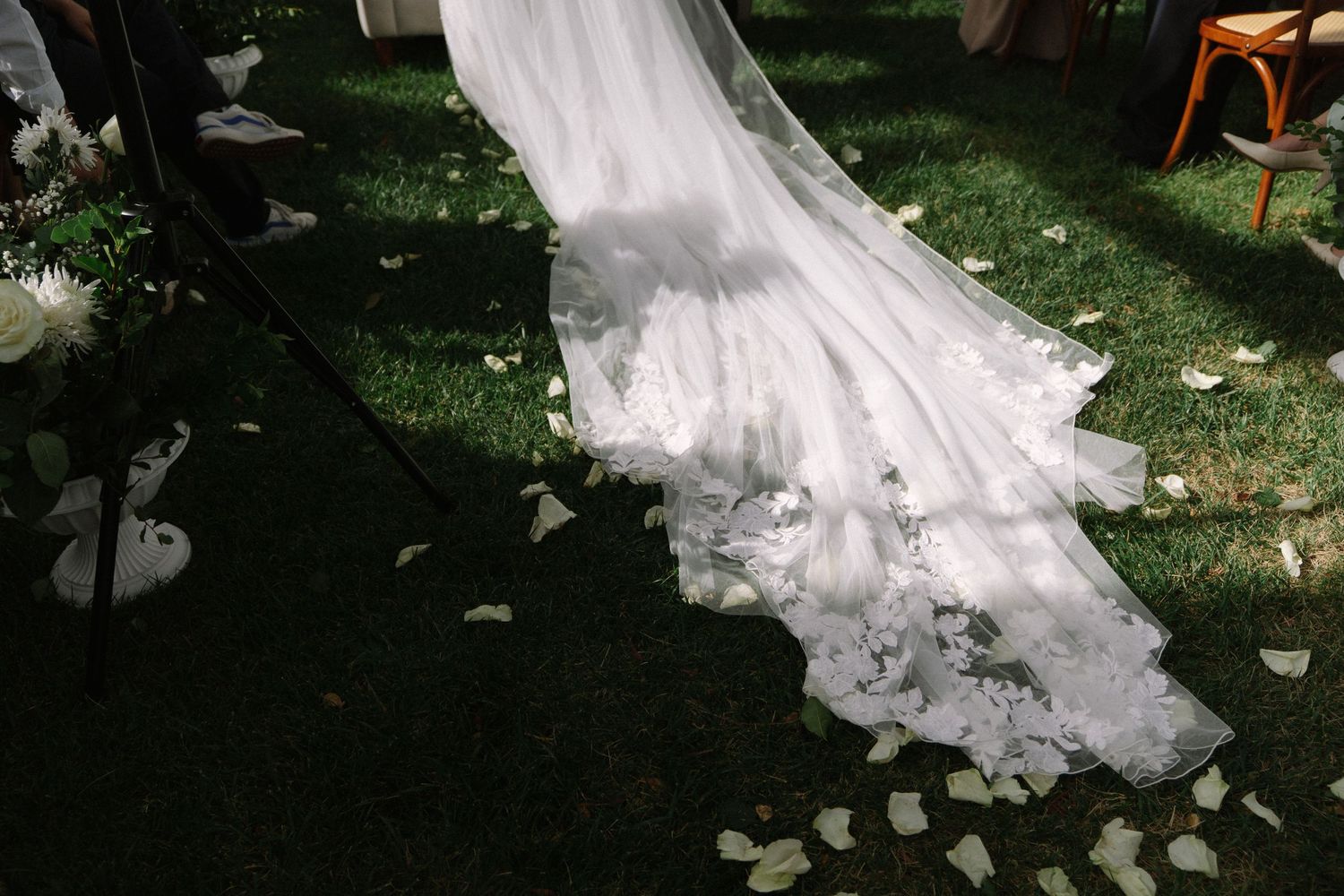 Elegant white lace wedding dress train spread on green grass with scattered white flower petals.