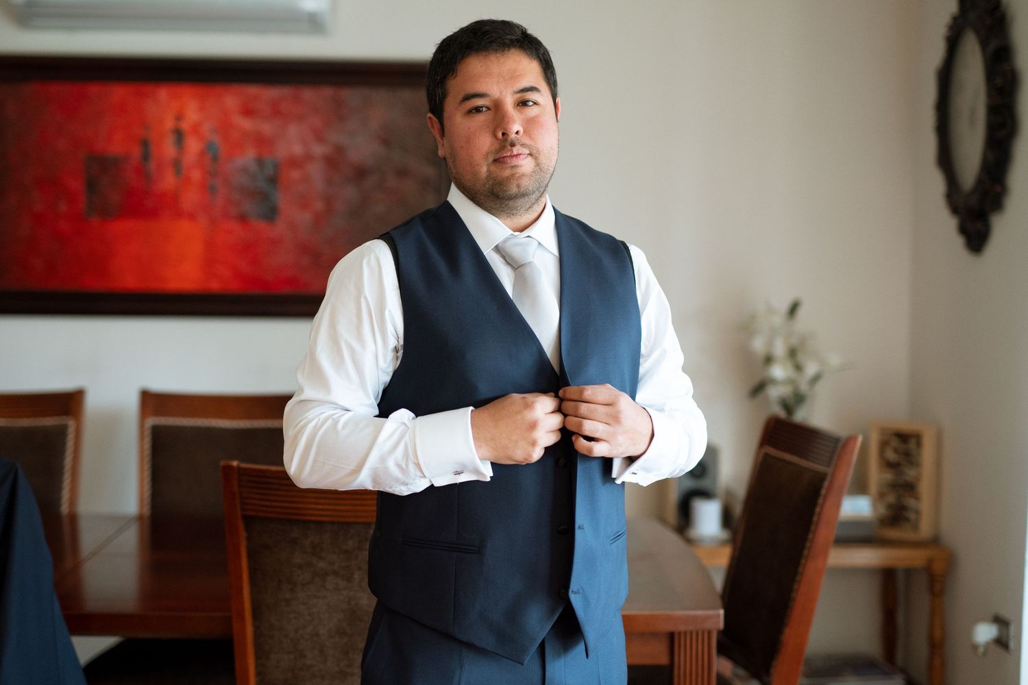 A groom adjusts his navy blue vest while getting ready in a dining room.