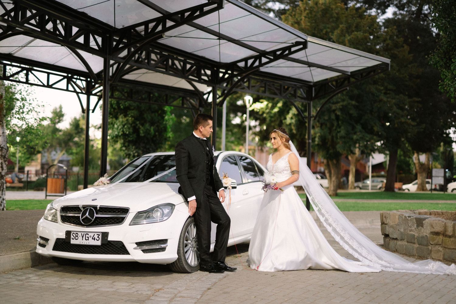 A wedding couple stands beside their luxury white Mercedes-Benz car under a metal canopy.