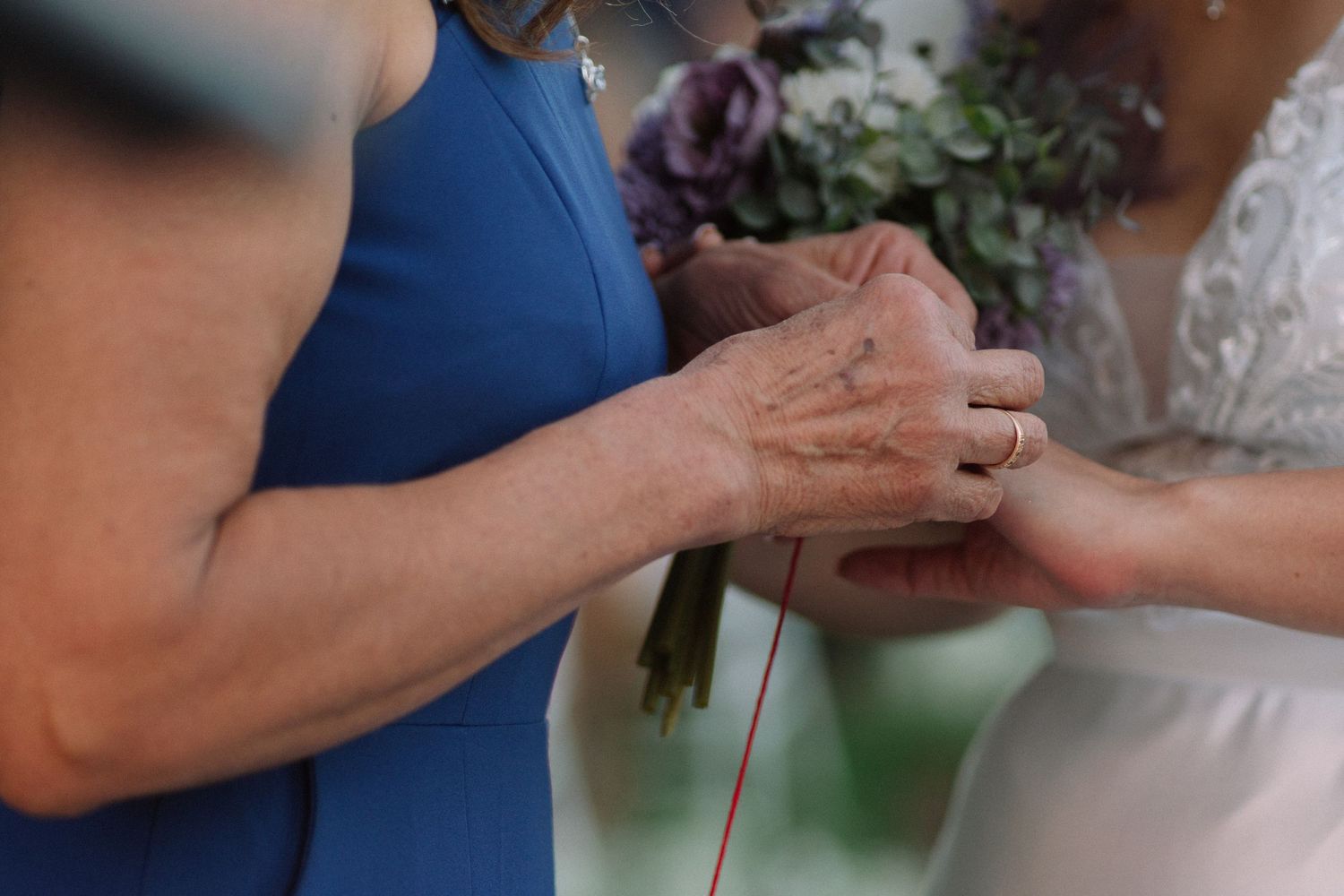 Close-up of hands exchanging rings with flowers visible in the background at outdoor wedding.