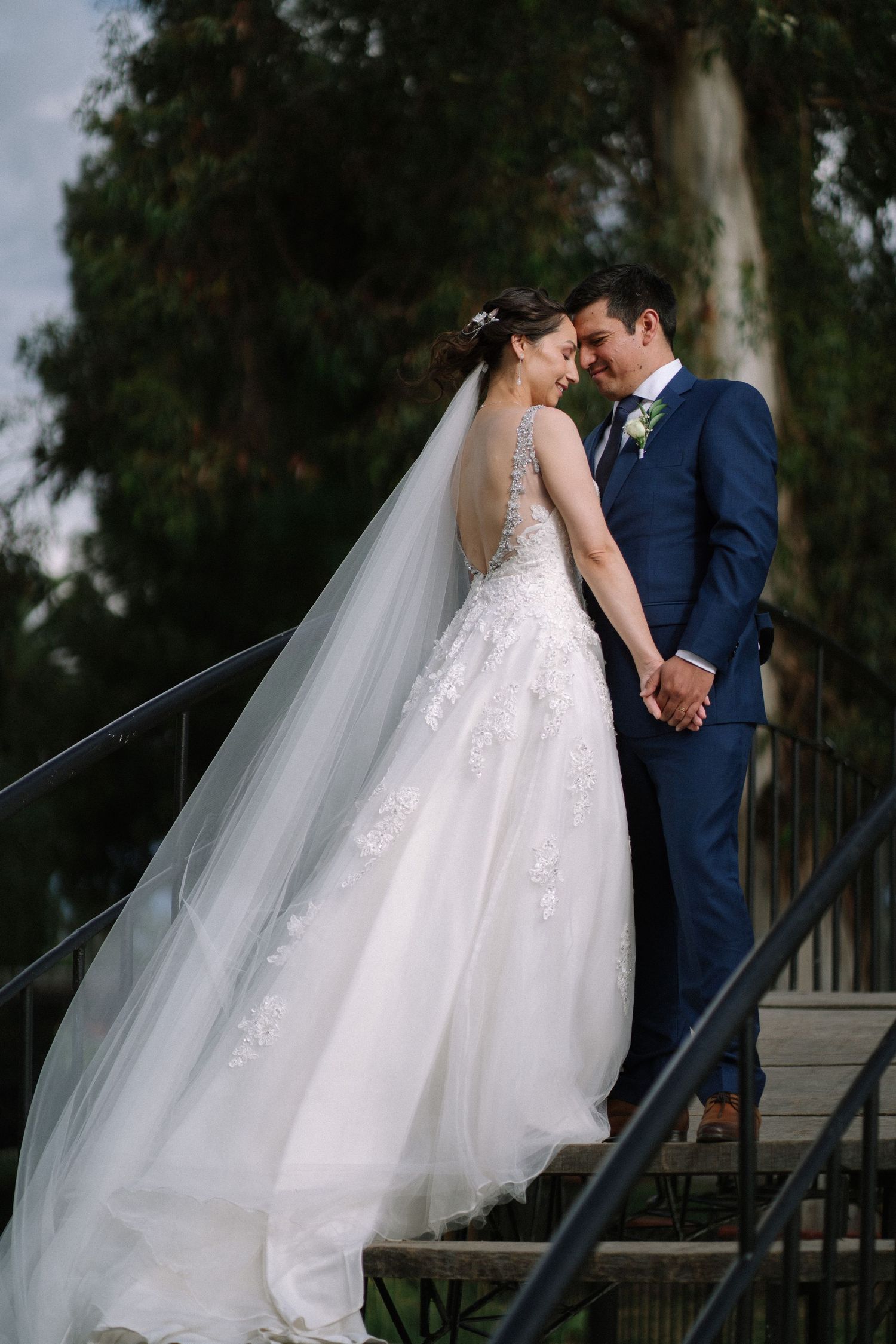 Couple shares tender moment on outdoor steps with bride in flowing lace gown and long veil.