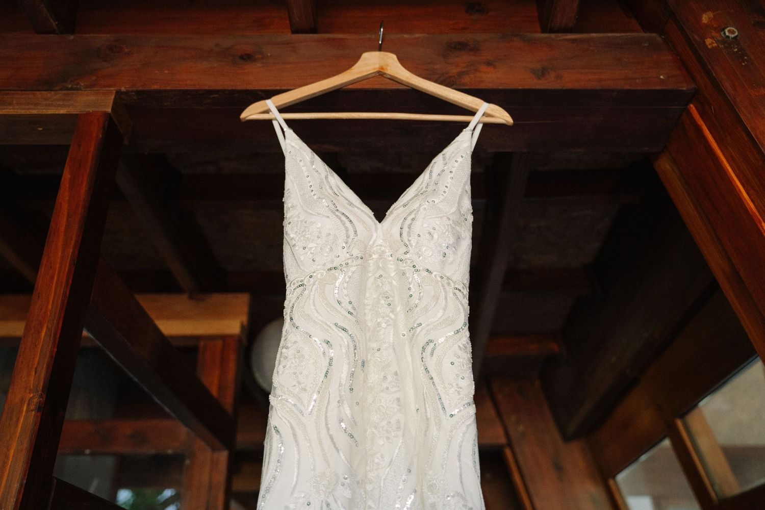 An elegant white lace wedding dress hangs on a wooden hanger against rustic beams.