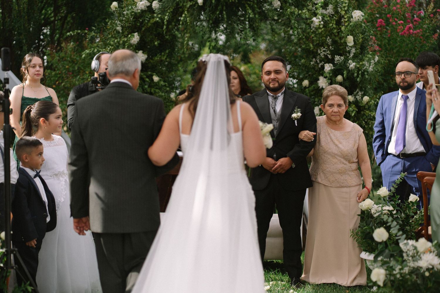 Bride in white gown with veil approaches groom at floral-decorated outdoor wedding ceremony altar.