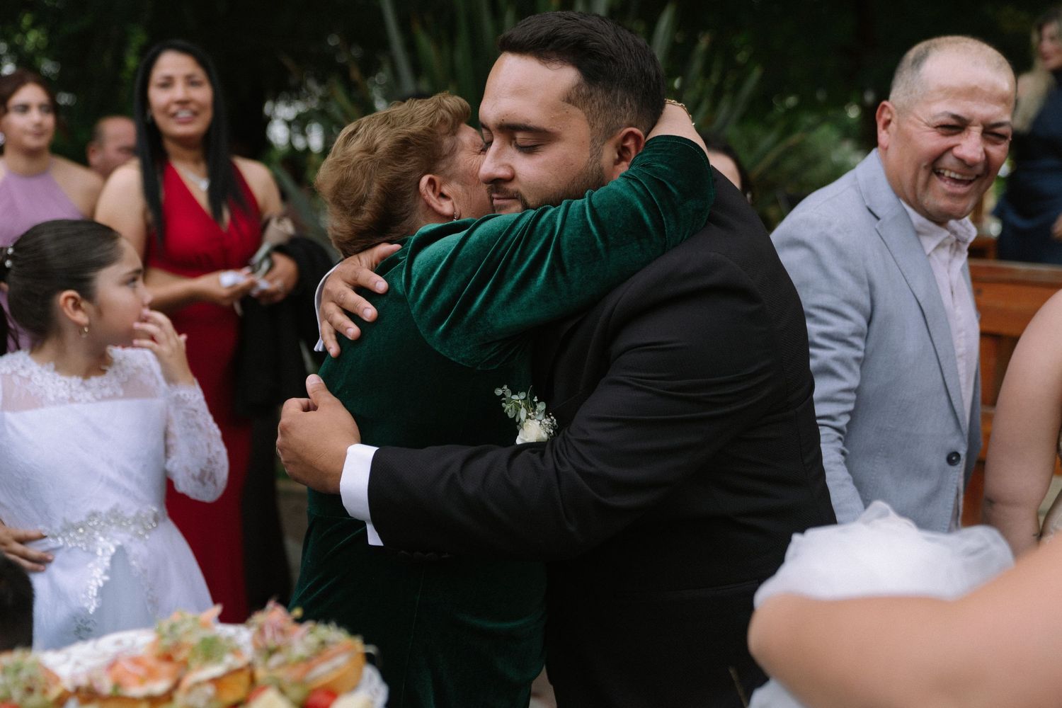 Groom in green velvet jacket hugging guest at outdoor wedding reception celebration.