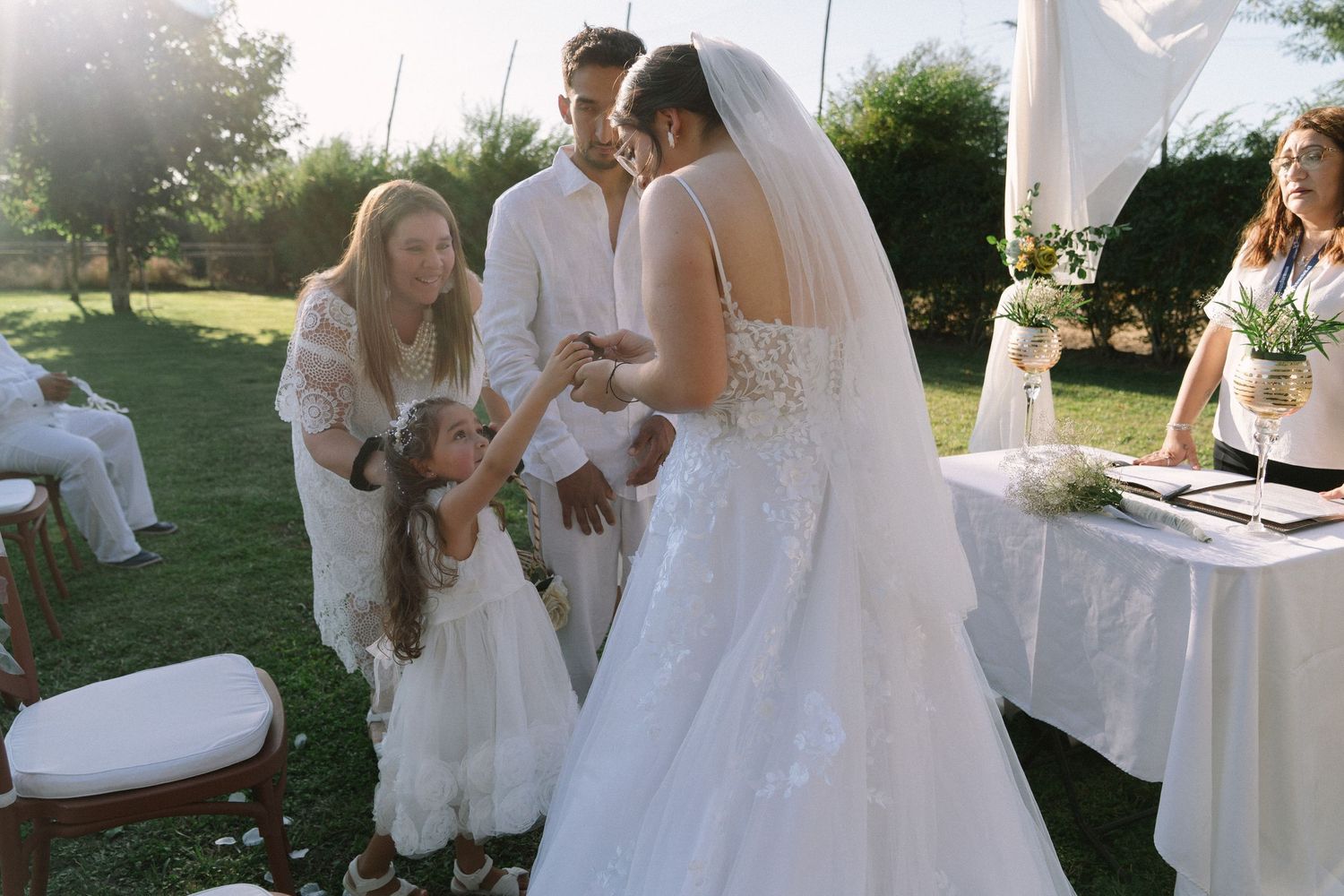 Outdoor wedding ceremony with bride in white gown, groom, flower girl, and officiant gathered on green lawn at sunset.