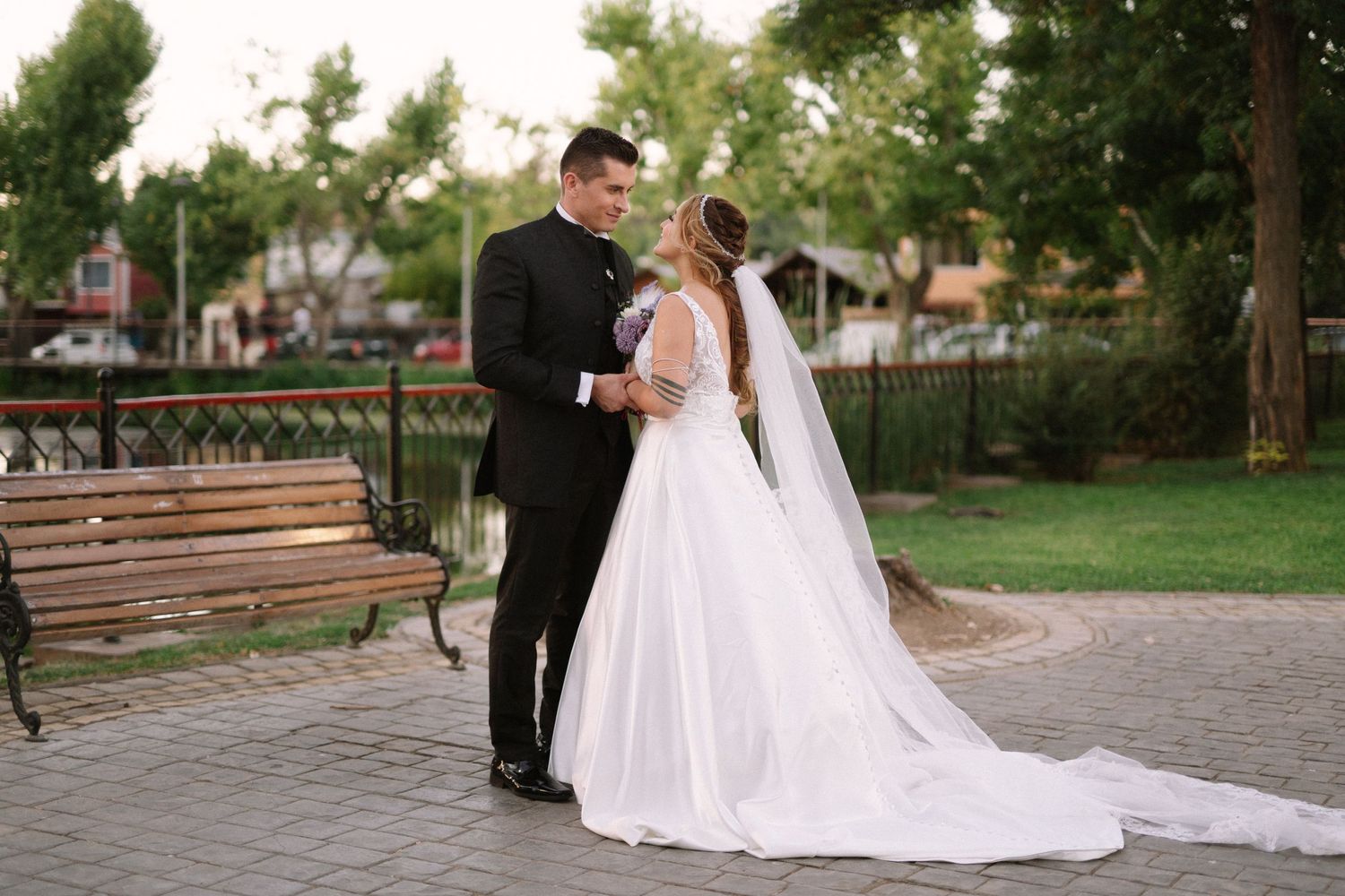 A wedding couple poses on a stone path with trees and a wooden bench in the background.
