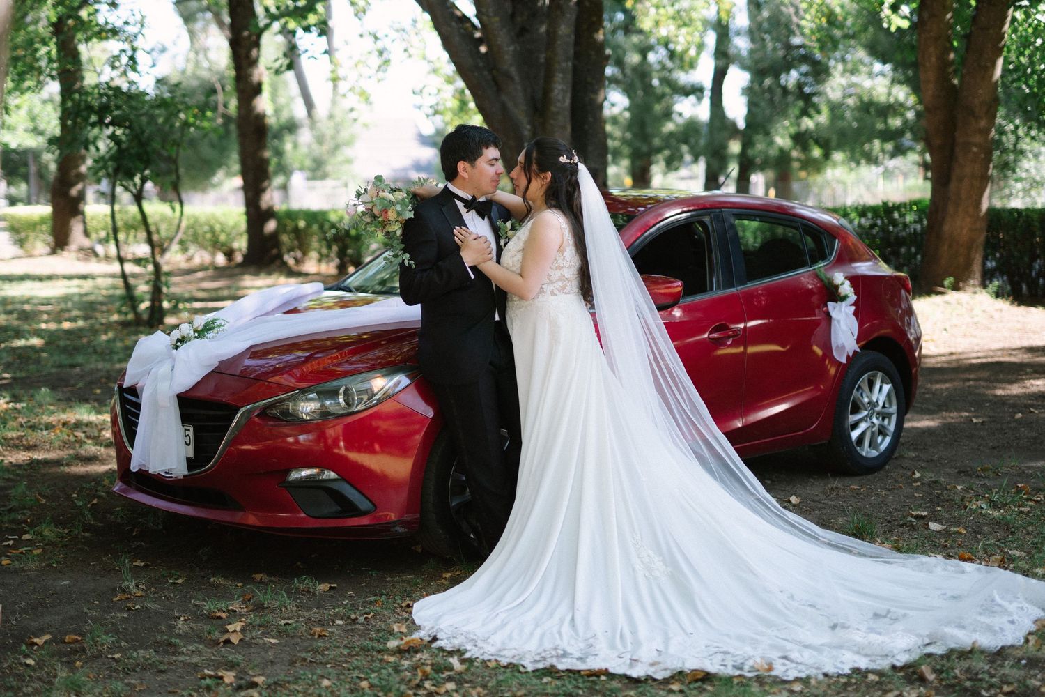 Wedding couple posing by red Mazda car under trees.