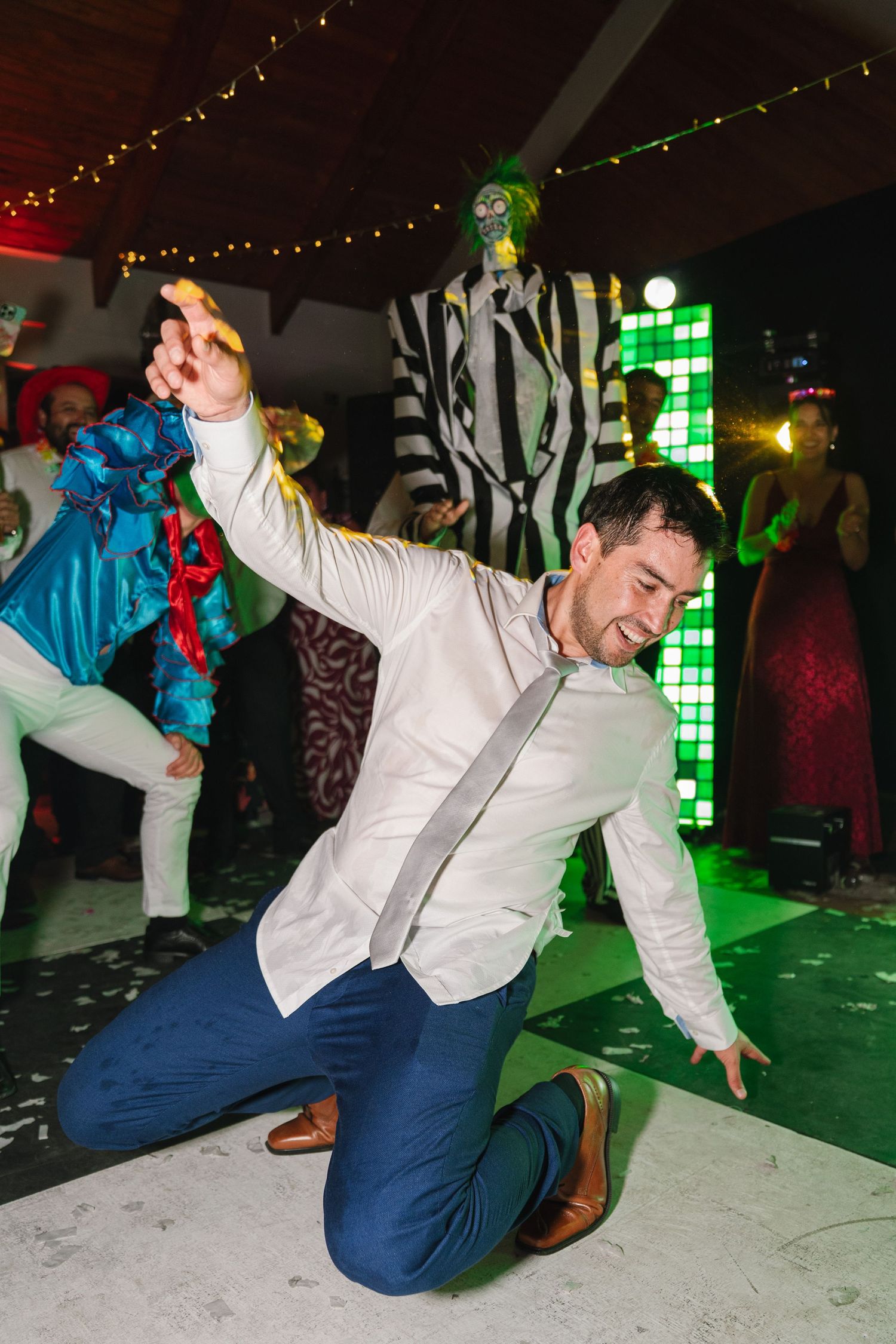 Wedding guest in blue pants and white shirt performs energetic dance moves on reception dance floor.