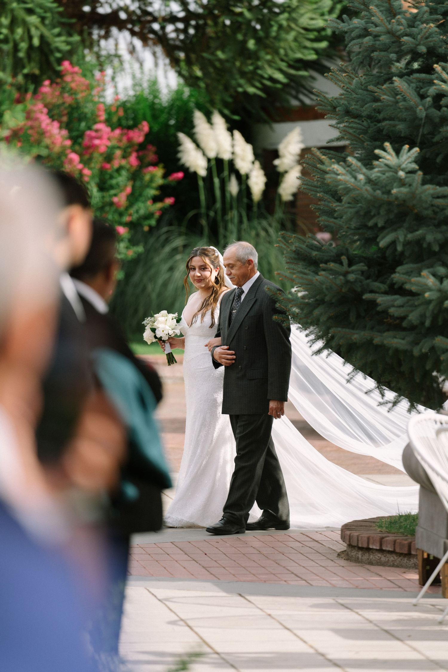Bride in white gown walks down outdoor aisle with escort amid lush greenery and floral arrangements.