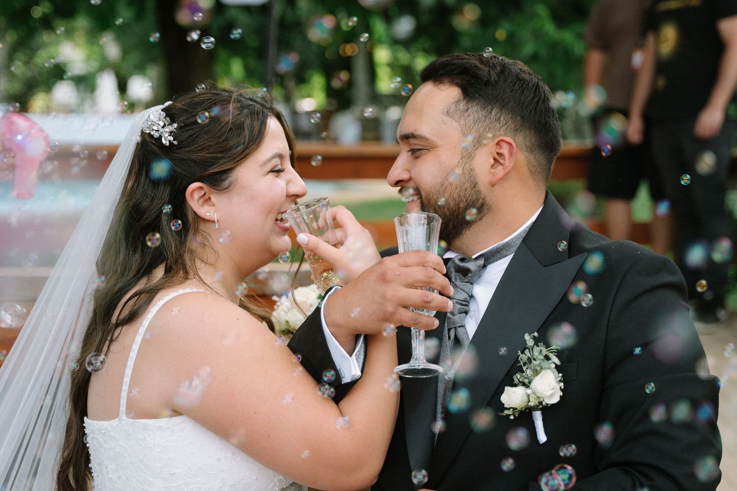 Bride and groom toast with champagne glasses surrounded by floating bubbles during joyful outdoor wedding celebration.
