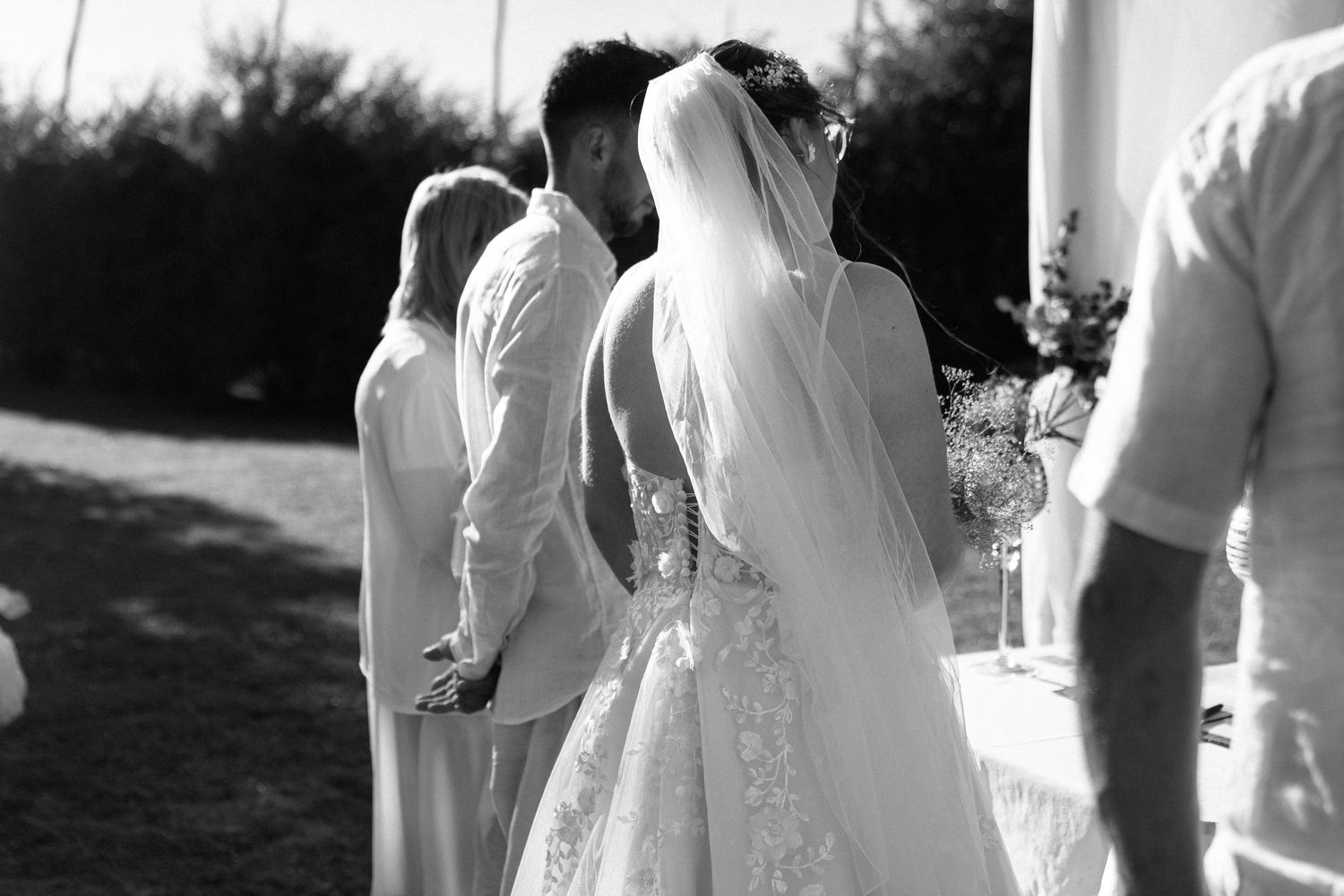 Black and white wedding photo of bride in lace gown and veil standing with groom during outdoor ceremony.