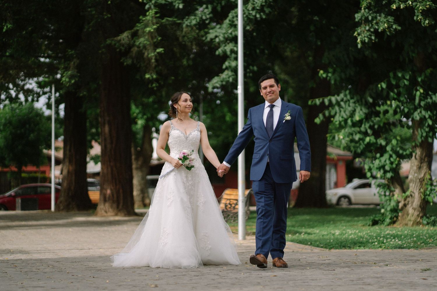 Newlyweds walk hand in hand on tree-lined path in formal attire.