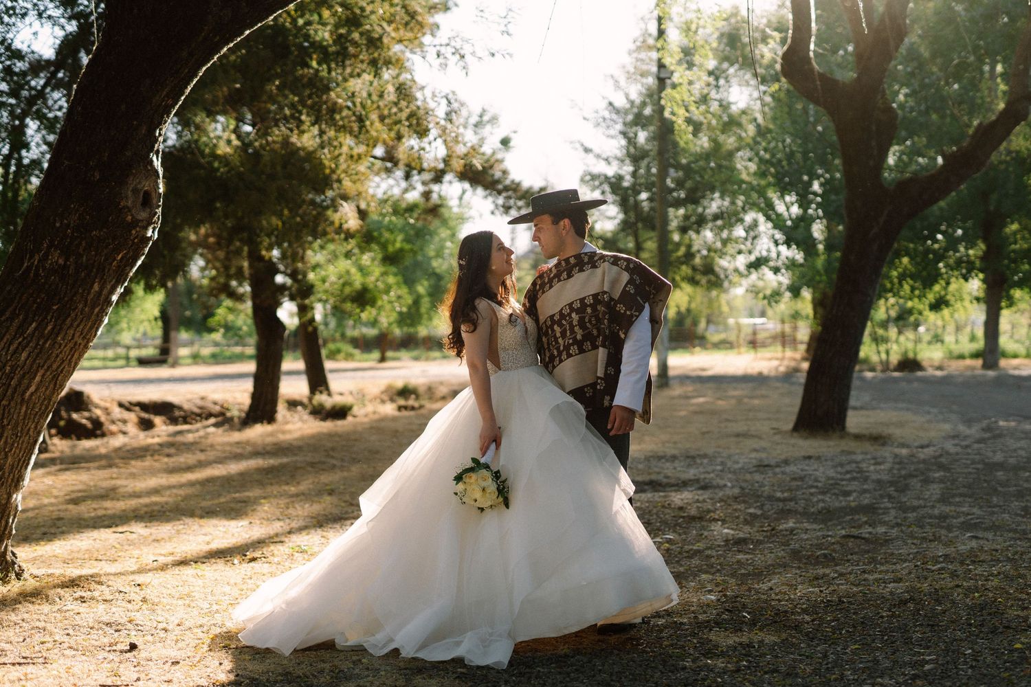 A romantic wedding photo captured in a sunlit wooded park setting with a flowing white gown and bouquet.