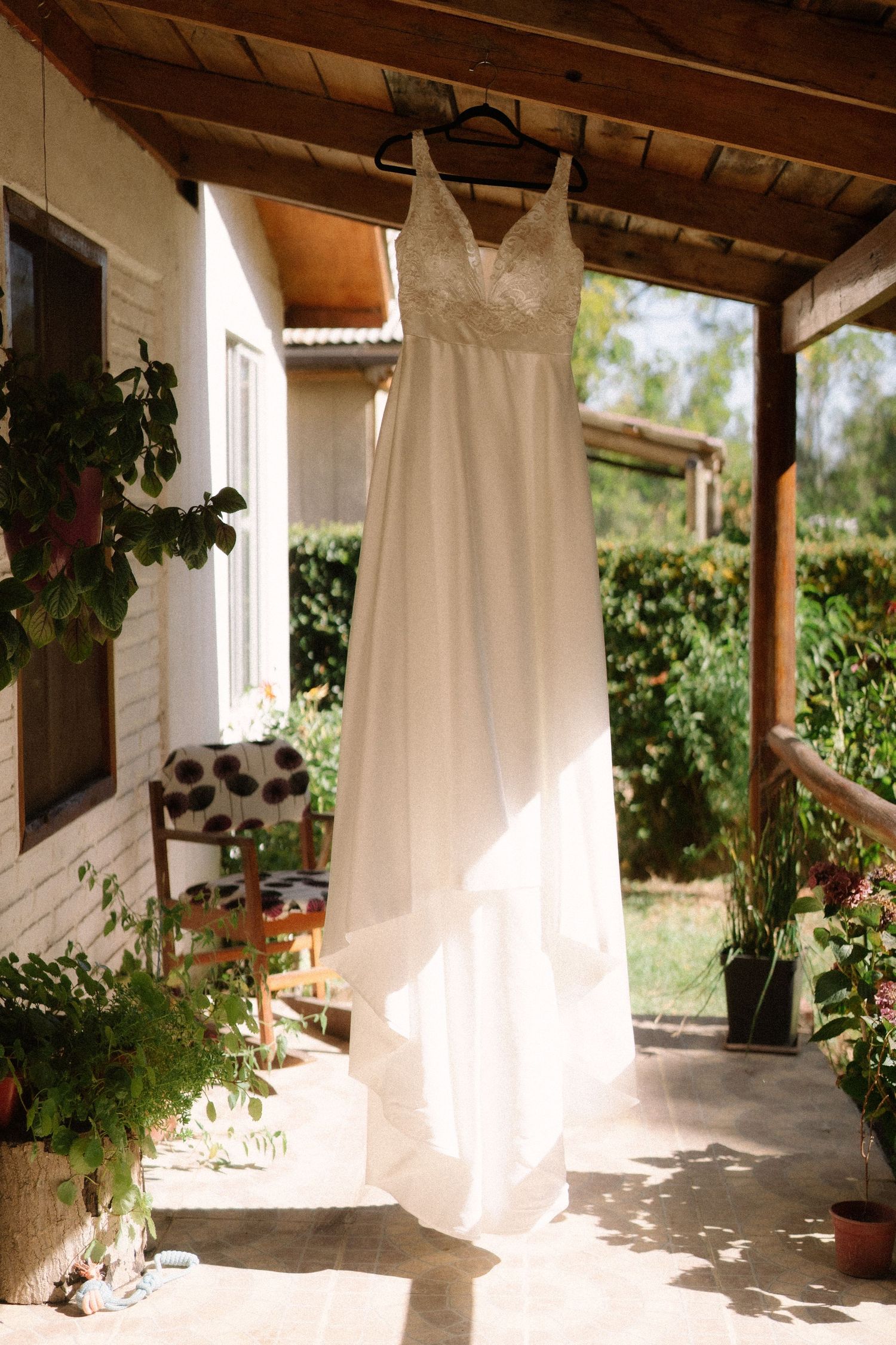 An elegant white wedding dress hangs on a porch with potted plants and natural lighting.