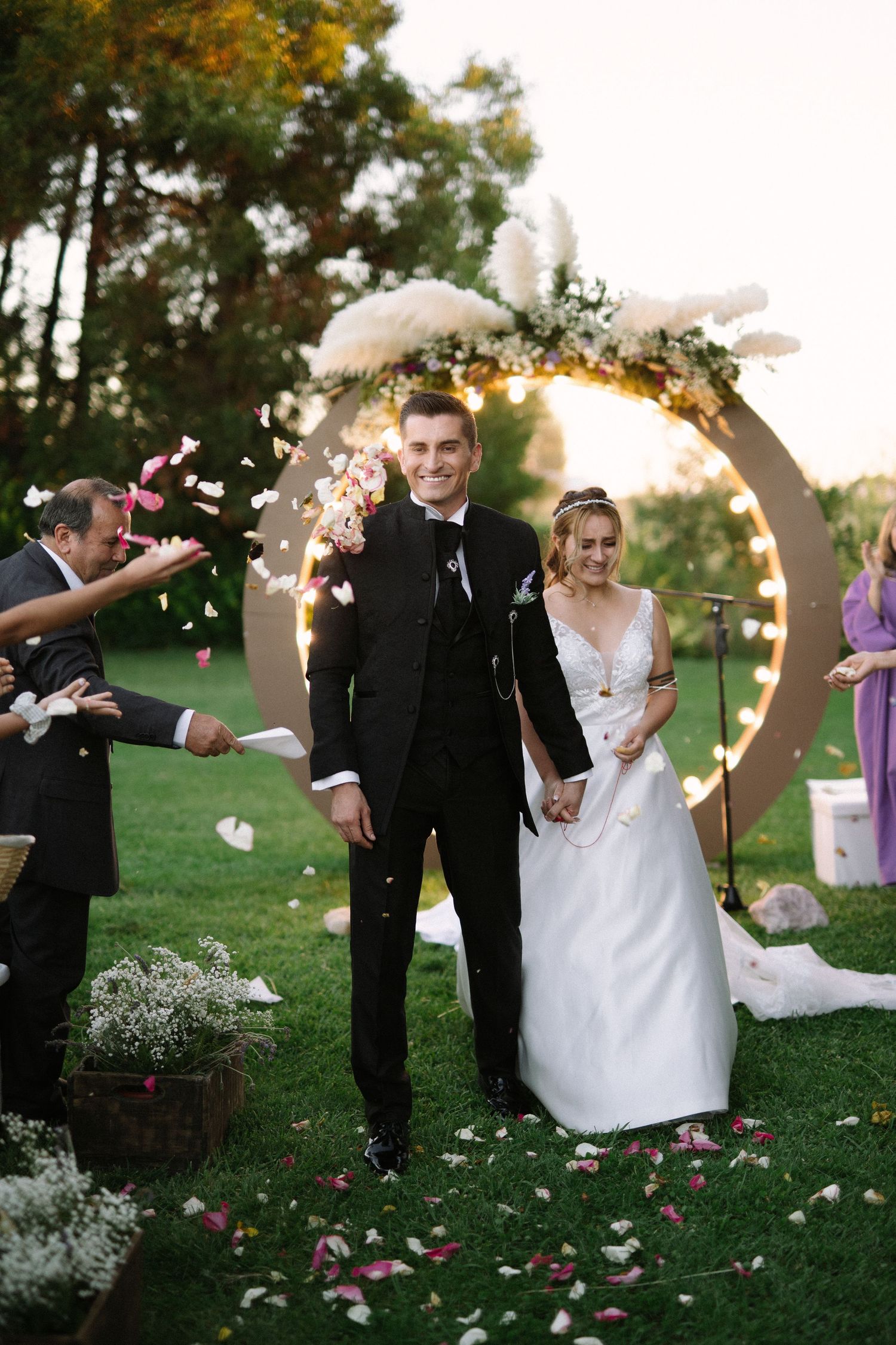 Guests shower newlyweds with pink rose petals under illuminated wedding arch at outdoor ceremony.