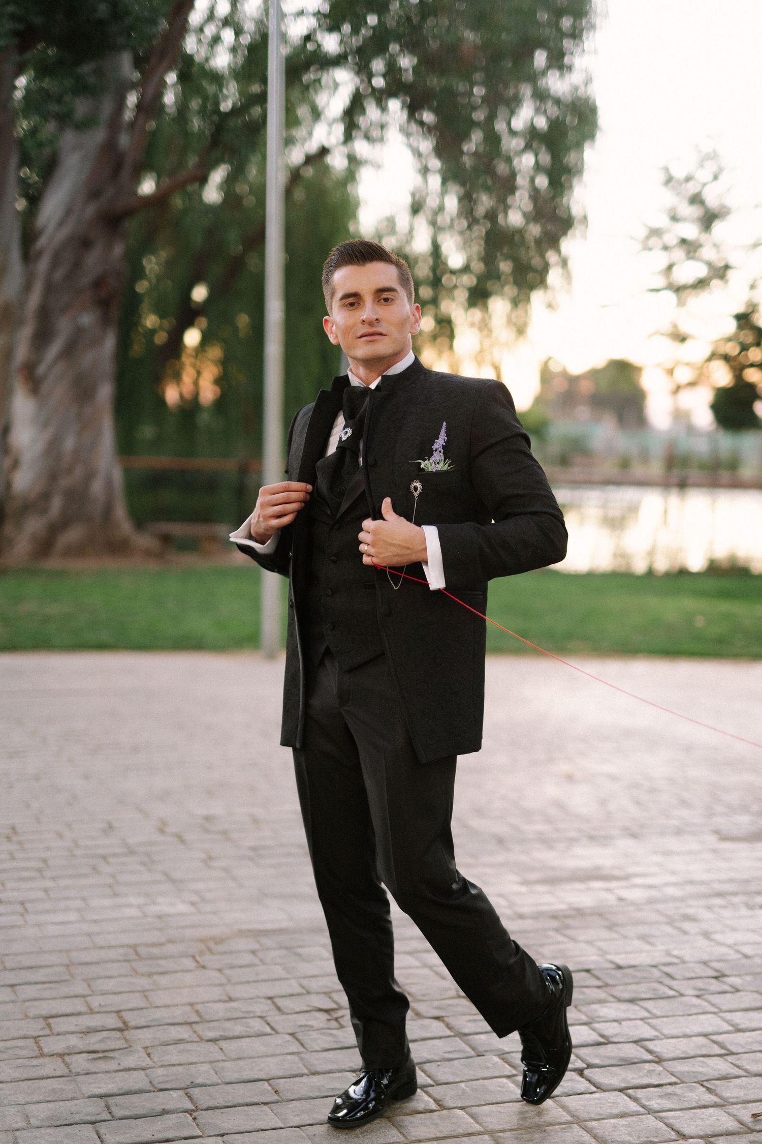 A groom stands confidently in a black tuxedo on a paved outdoor path at sunset.