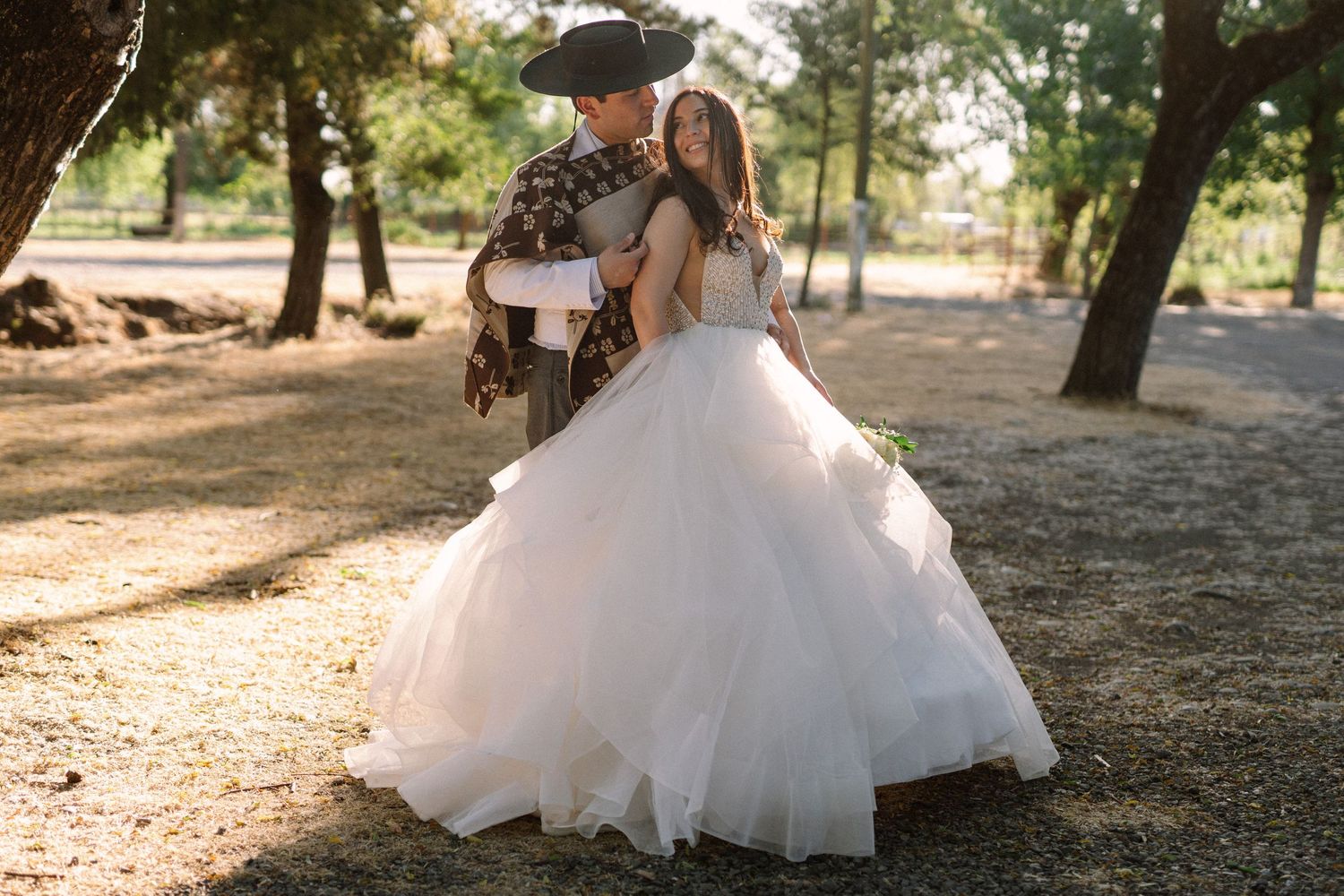 Wedding couple embraces on sunlit path surrounded by trees.
