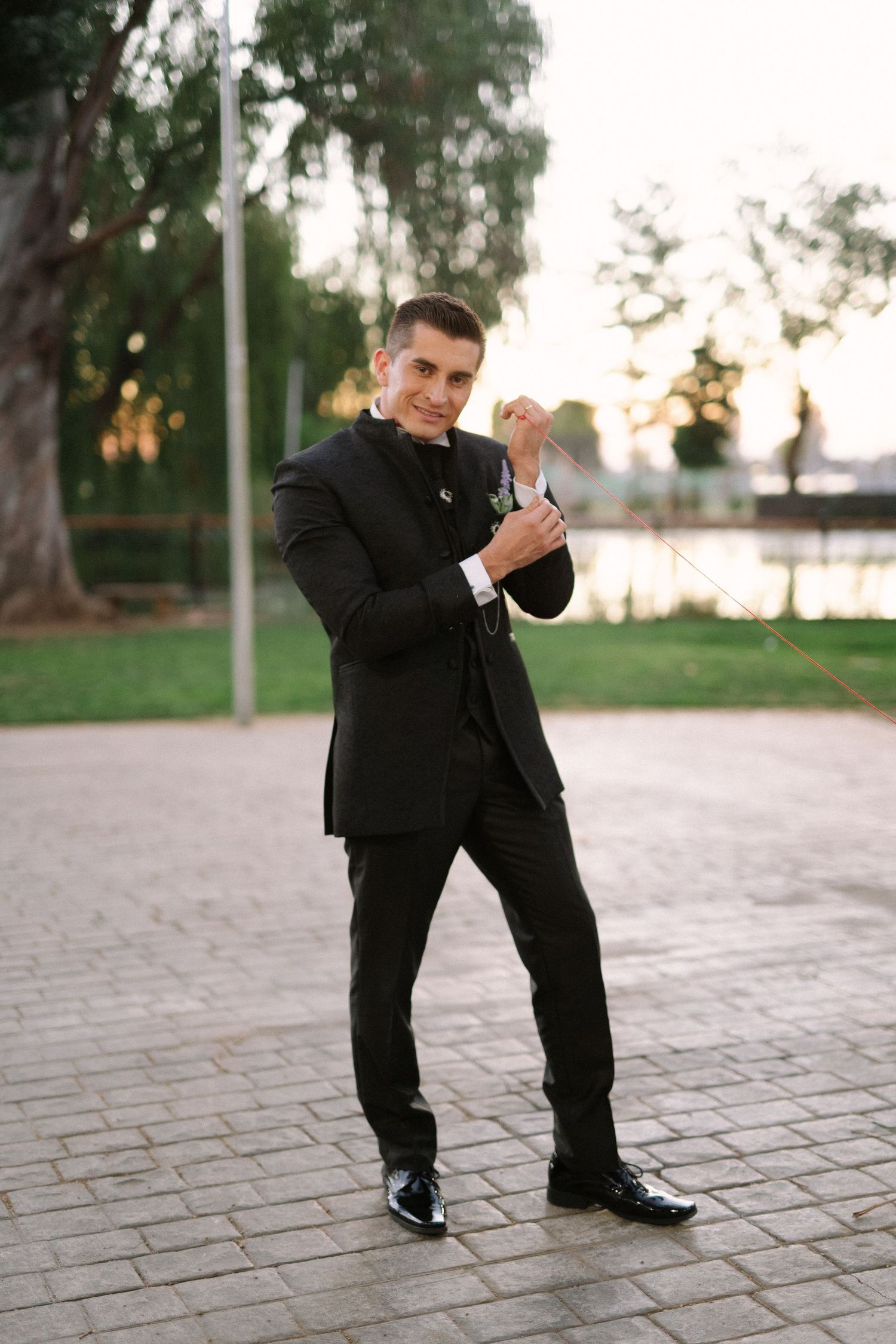 A groom in classic black tuxedo adjusts his cufflinks while posing on stone pavers outdoors.