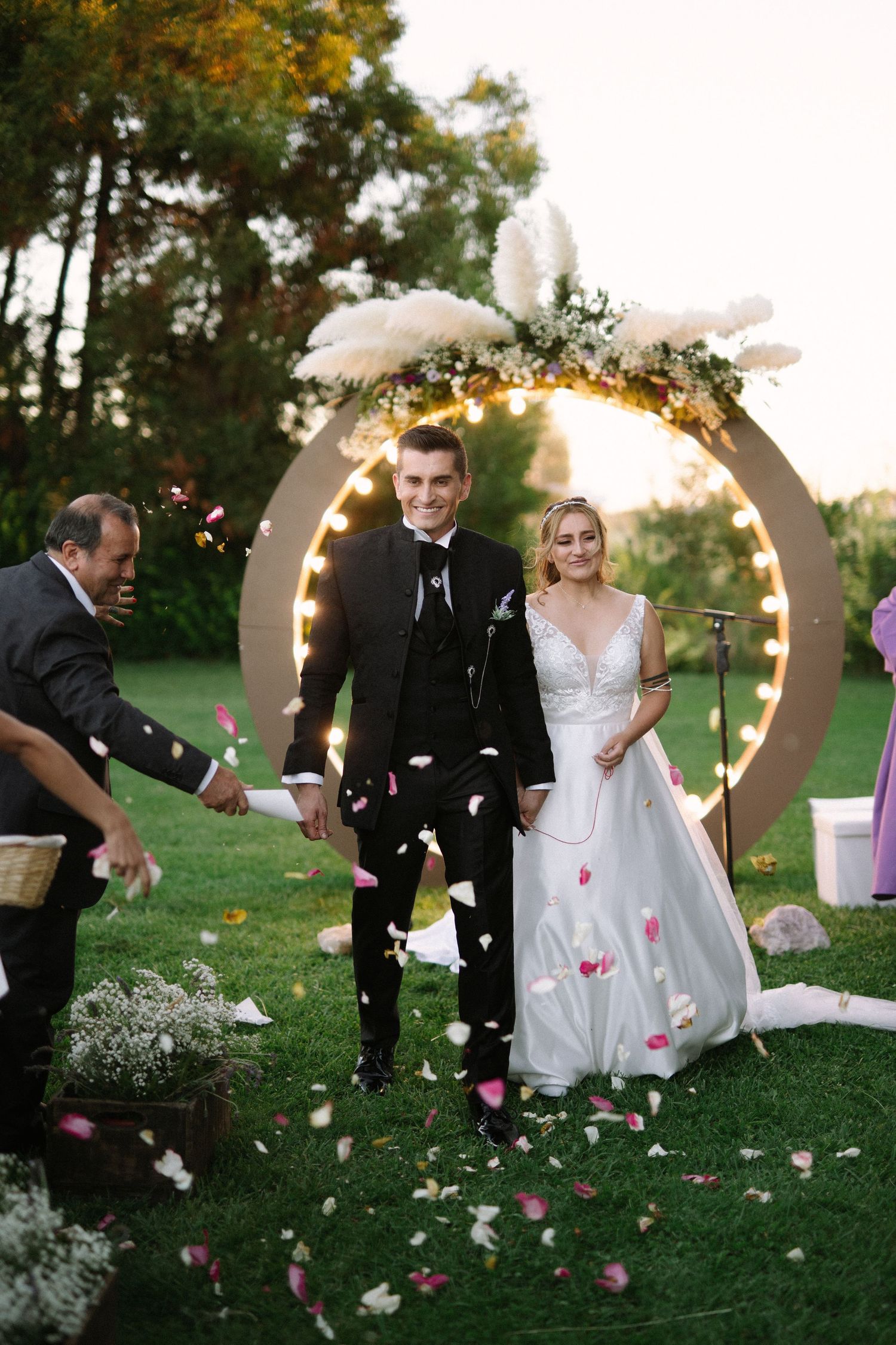 Wedding guests toss rose petals at couple under glowing circular arch with feather decor.