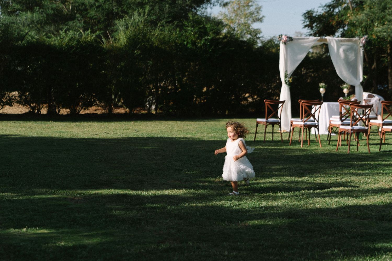 A flower girl in a white dress runs across a lush green lawn at an outdoor wedding ceremony.