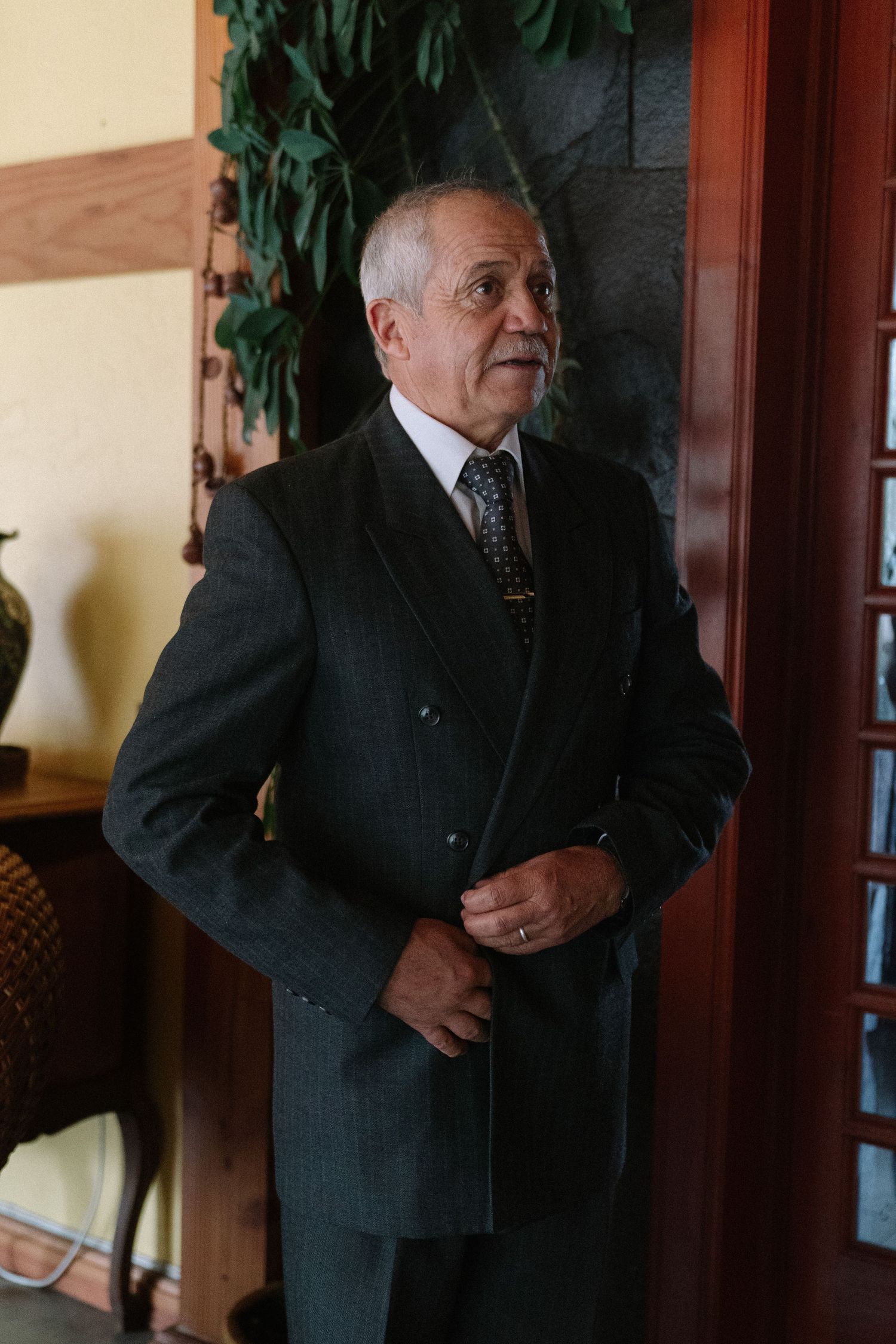 Elegant older man in dark double-breasted suit adjusting jacket indoors near wooden door.