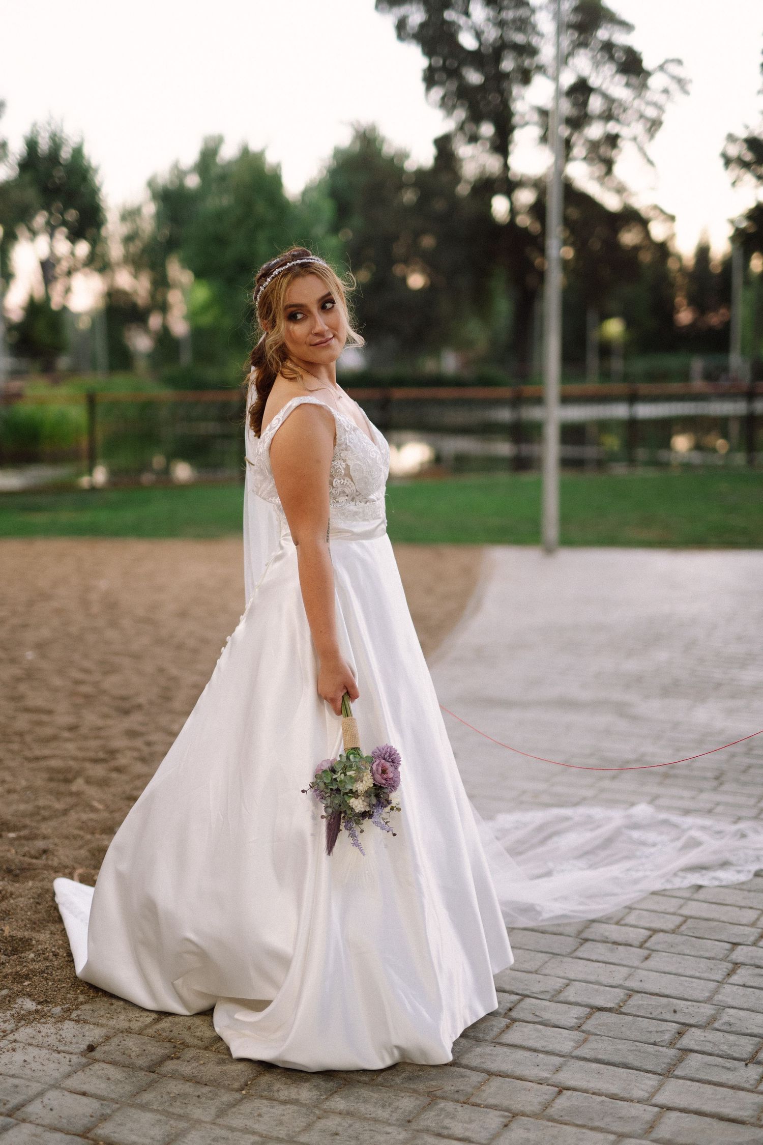 A bride in an elegant white gown holds a purple bouquet while posing outdoors.