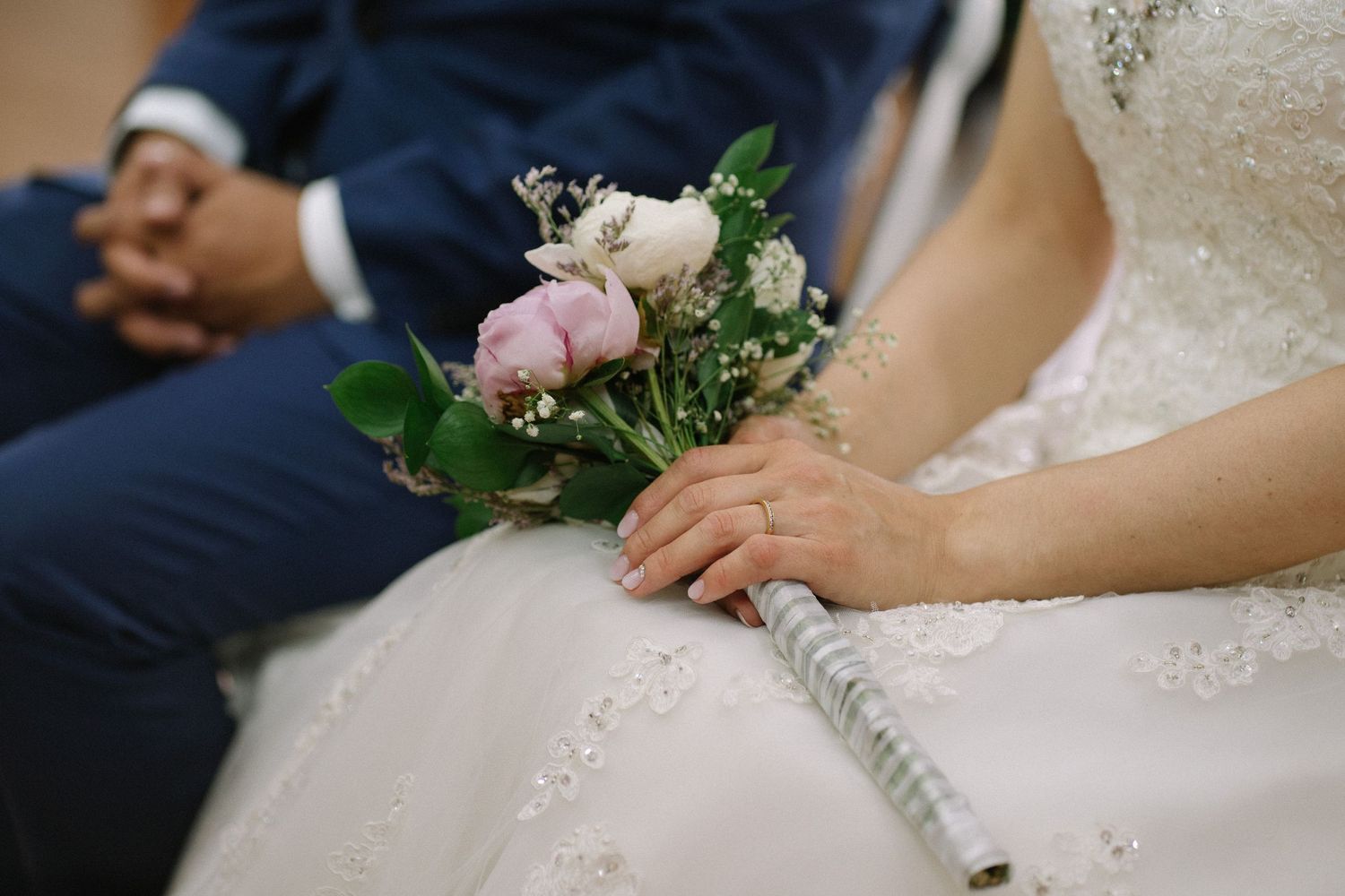 Wedding bouquet with pink roses resting on white dress with navy suit in background.