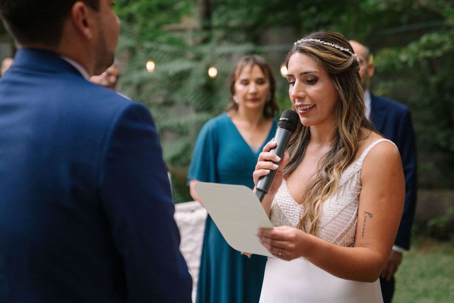 A bride reads from paper during an outdoor wedding ceremony while wearing a white lace dress.