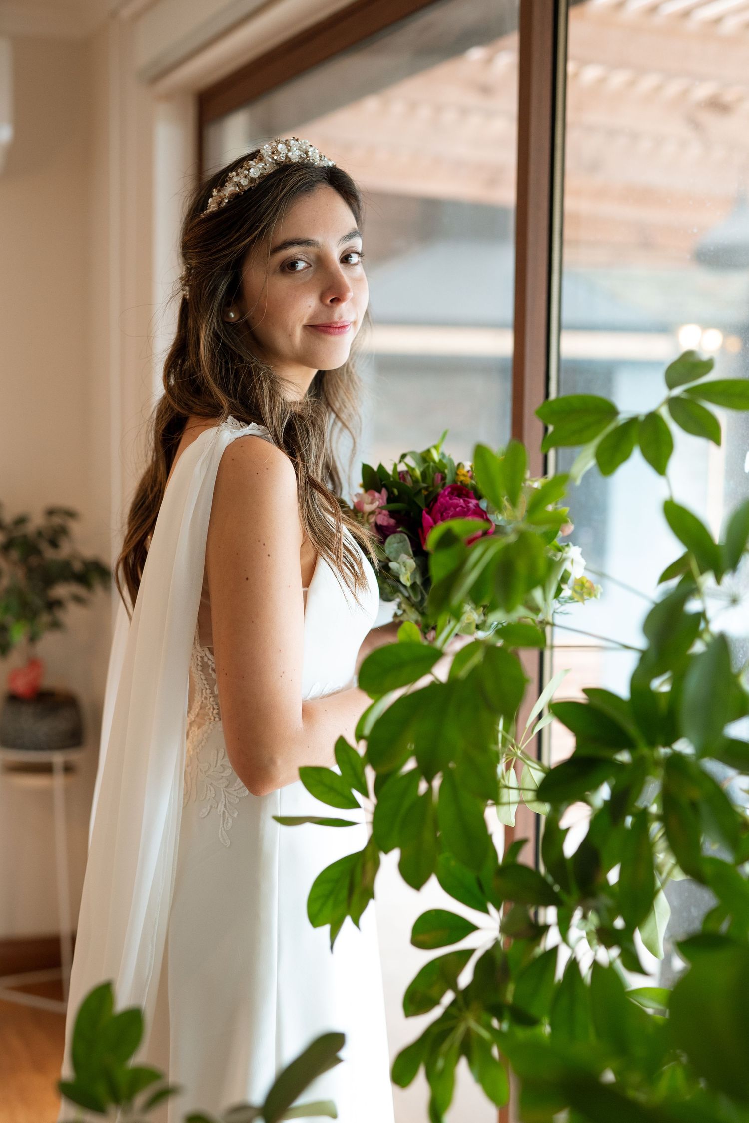 A bride in an elegant white gown poses near a window with green foliage in the foreground.