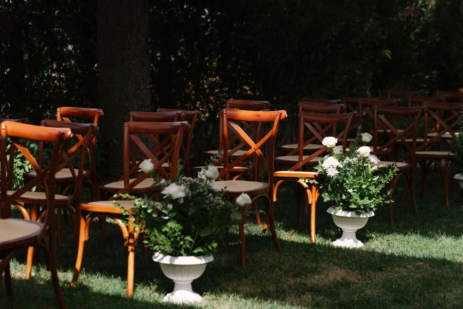 Wooden cross-back chairs arranged in rows outdoors for a wedding ceremony with white floral arrangements.
