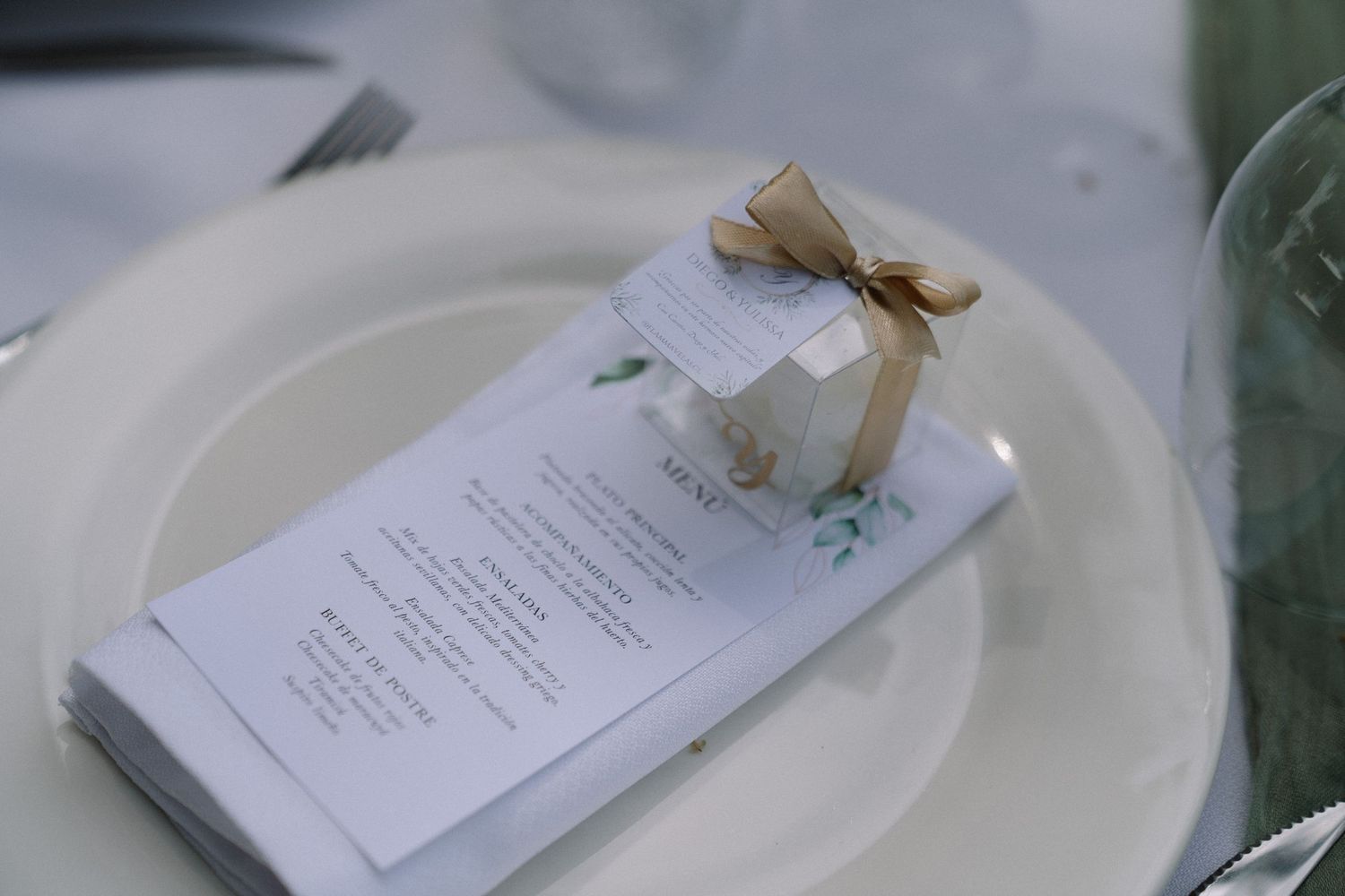Elegant wedding place setting with white plate, menu card, and small gift box tied with gold ribbon.