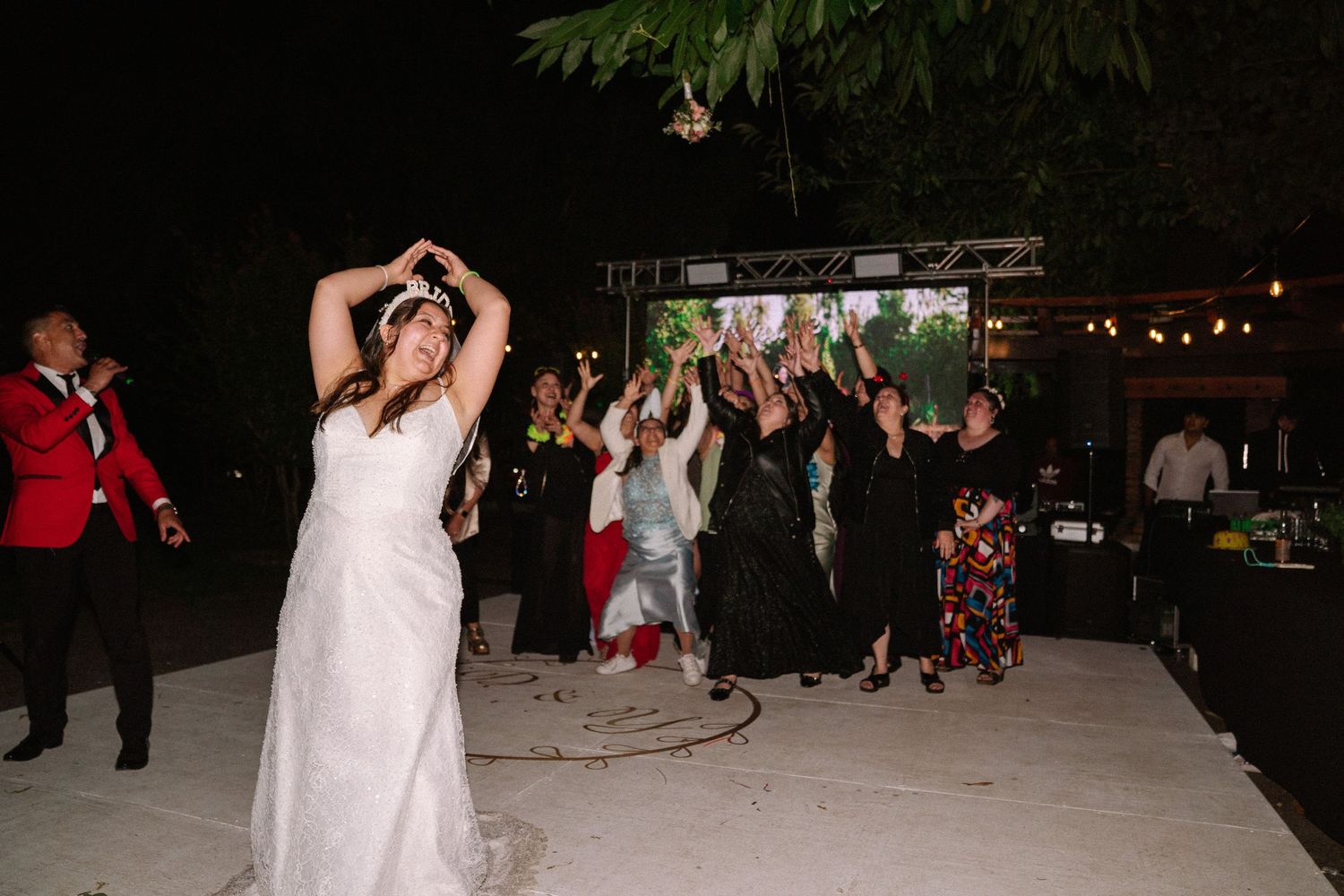 Bride tossing bouquet to excited guests on outdoor dance floor during wedding reception.