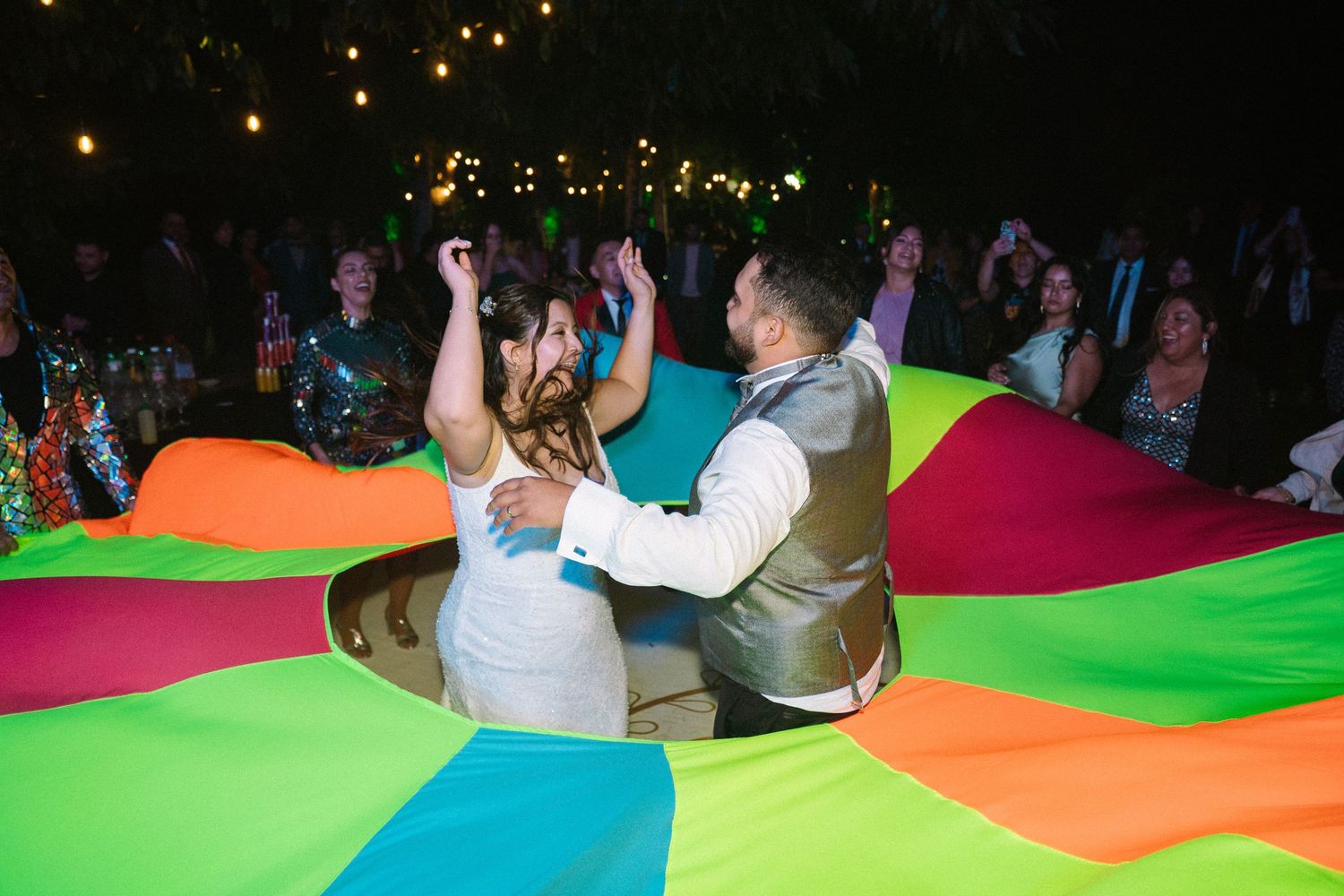 Bride and groom dance inside colorful parachute at lively outdoor wedding reception at night.