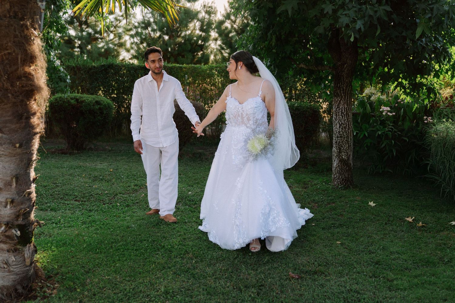 Bride and groom hold hands walking through lush green garden on their wedding day.