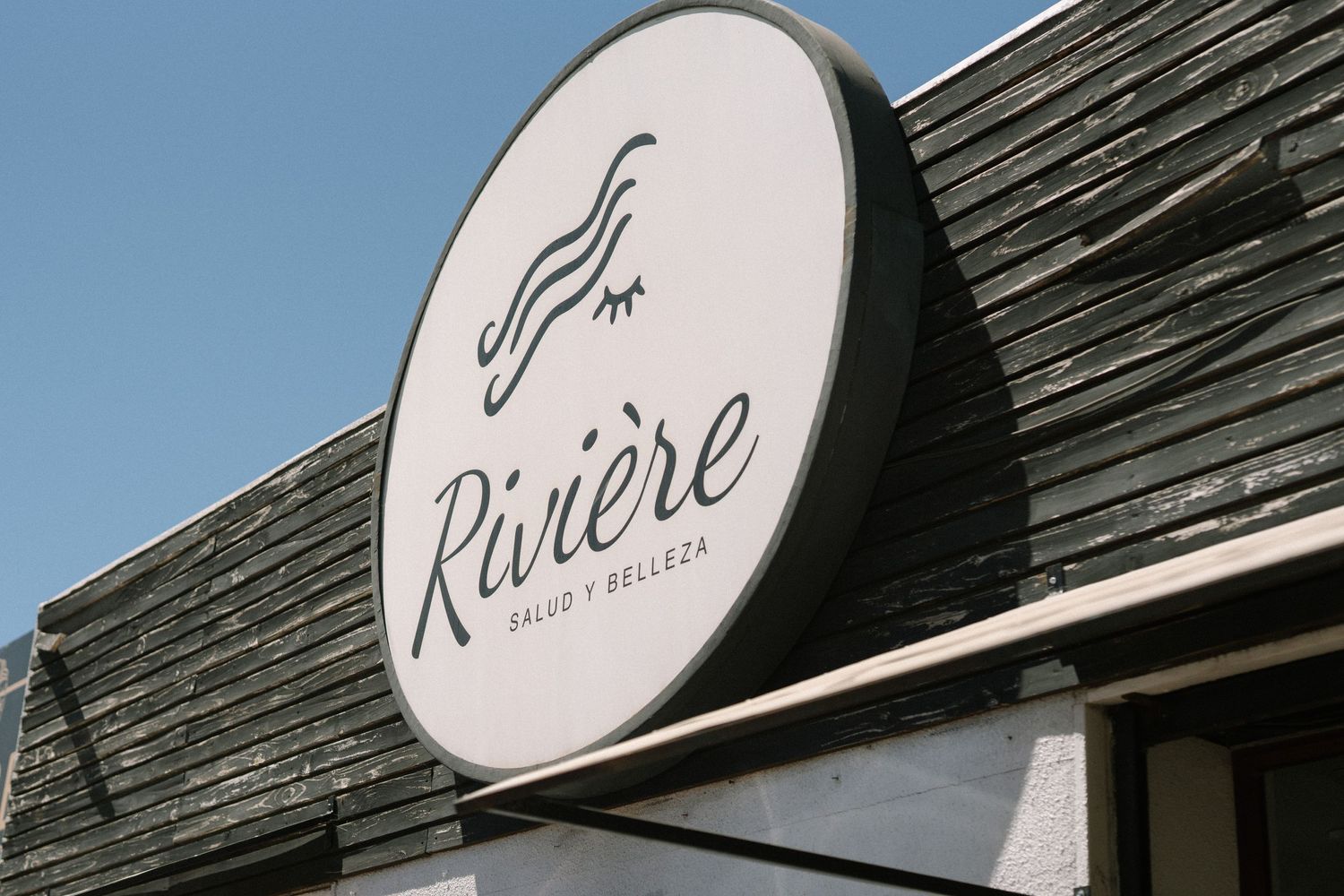 Riviere salon and boutique sign mounted on a dark shingled roof against blue sky.