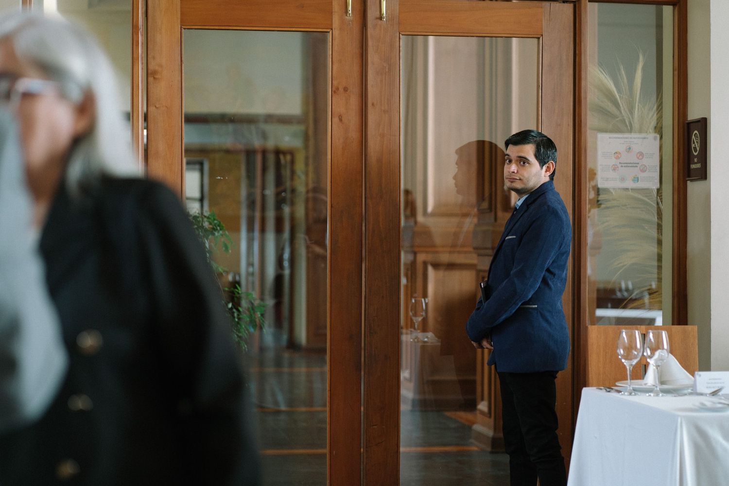 A person in a navy suit stands near elegant wooden doors in an event space.